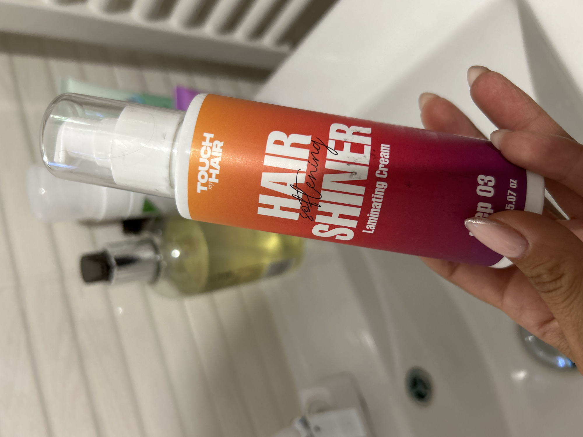 A hand with light pink nails holds a "TOUCH my HAIR" brand "HAIR SOFTENING SHINER Laminating Cream" bottle. The bottle has an orange to pink gradient and white text. Behind the bottle, a sink with a faucet is visible, along with other toiletries, including a clear bottle with yellow liquid and a white pump bottle. The scene appears to be indoors, possibly in a bathroom, with a wall that has horizontal lines. There is no indication of the city of Burgas, Bulgaria, in the image.