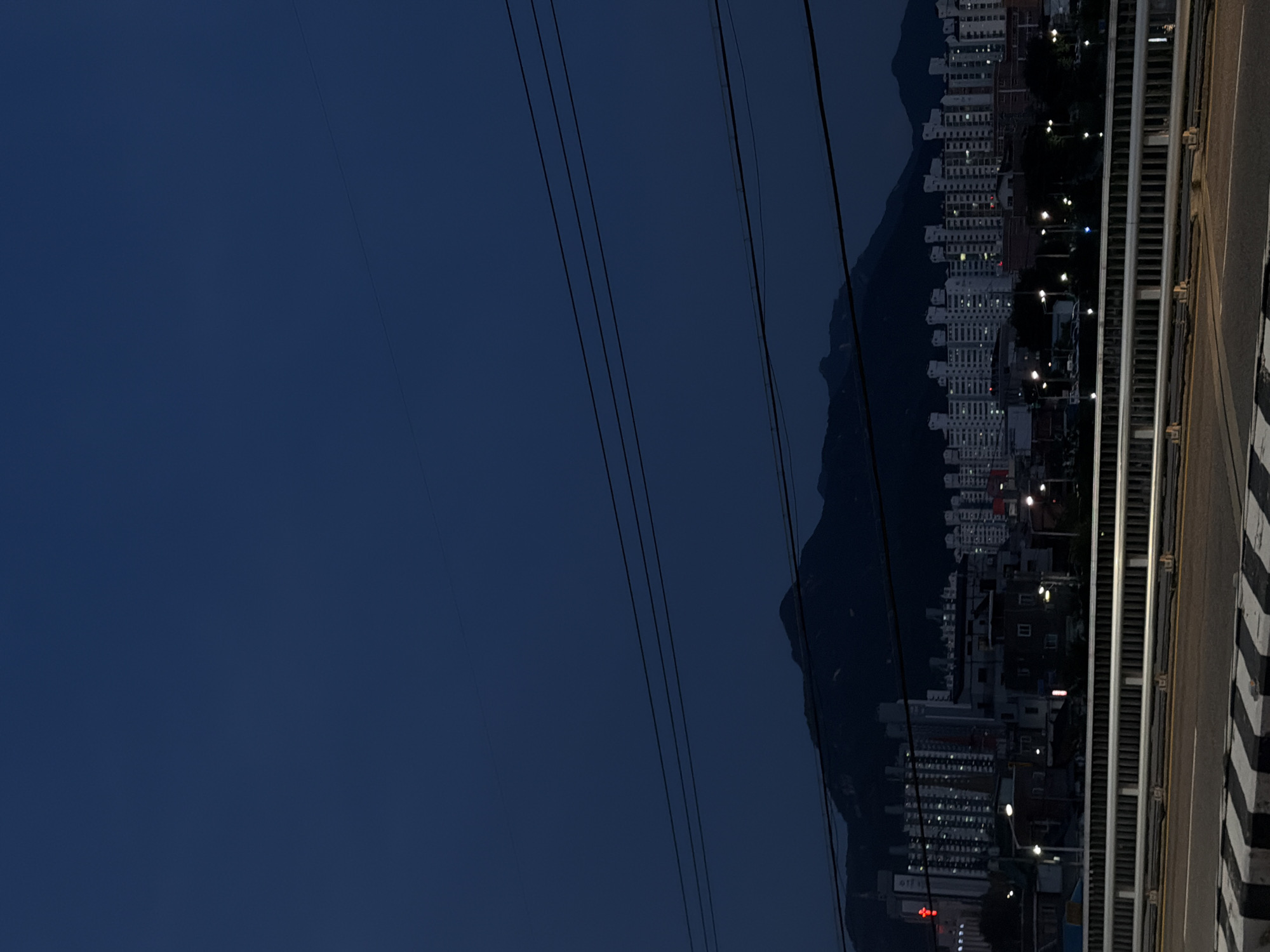The image shows a view of Uijeongbu-si, South Korea, at dusk. The foreground features a bridge with a metal railing and a road marked with yellow and black stripes. Beyond the bridge, a cityscape unfolds with numerous apartment buildings and some smaller structures. Streetlights are visible, illuminating parts of the urban landscape. In the background, dark, silhouetted mountains rise against the deep blue twilight sky. Power lines are strung across the upper portion of the image, adding a linear element to the composition. No people are clearly visible in the image. The overall scene depicts a city transition from day to night, with the urban environment set against a natural mountainous backdrop.