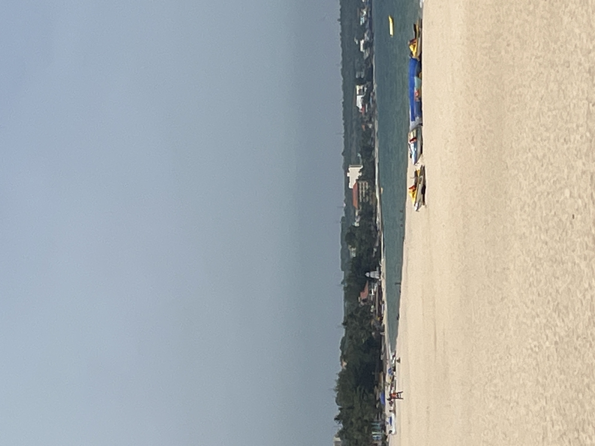 The image depicts a wide expanse of sandy beach in Mũi Né, Vietnam, with the ocean stretching out towards a hazy horizon. The sand is a light beige color and appears mostly unoccupied, with a few scattered beach chairs and umbrellas visible in the distance. Closer to the water's edge, several watercraft are parked on the sand. These include at least two jet skis, a blue inflatable raft, and what appears to be another watercraft covered in a blue tarp. In the background, a line of lush green trees forms the coastline, behind which are buildings of varying sizes and colors, suggesting a developed area. A prominent white lighthouse-like structure stands out amongst the buildings. The sky is a uniform light gray, indicating an overcast or cloudy day. There are no prominent actions or interactions captured; the scene is one of quiet repose.