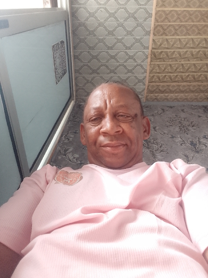 An adult male with dark skin and a bald head is the primary subject of this close-up, upward-angled selfie. He appears to be middle-aged or older, with visible wrinkles on his forehead and around his eyes, and a relaxed, slightly smiling expression. He is wearing a light pink, ribbed, long-sleeved shirt. A small, stylized emblem or patch in shades of orange and brown, possibly depicting an abstract design or an animal head, is visible on the left side of his chest.
The setting is indoors, featuring a varied background of patterned walls and a window or door. To the upper left, a light-colored (possibly white or light grey) frame of a window or door is visible. Within the glass pane of this structure, a dark, rectangular sticker or label with small, dense, illegible text or symbols is affixed.
The wall behind the man is adorned with at least three distinct patterns. The upper left section, behind his head, features light grey wallpaper or tiles with a repeating wavy, interlocking geometric design. To the right of this, possibly separated by a light wooden strip, is a section of brownish-beige wallpaper or tiles displaying a different, more angular and geometric pattern, which might have a three-dimensional effect. Below these upper patterns, closer to the bottom right of the frame, the wall is covered with a darker grey wallpaper featuring a large, swirling floral or damask-like motif. The lighting appears soft and even, likely natural light illuminating the scene.