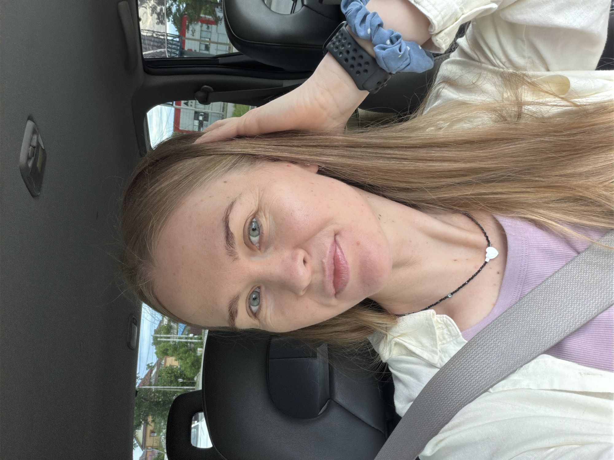 A Caucasian woman with blonde hair and blue eyes is taking a selfie in the driver's seat of a vehicle. She is wearing a light-colored shirt over a purple top, a necklace with a heart pendant, and a smartwatch with a blue scrunchie on her wrist. Her hand is resting on her head. The interior of the car is visible, including a black headrest and a grey seatbelt. Outside the car windows, buildings and trees can be seen, suggesting an urban or suburban environment. The scene appears to be set in Khabarovsk, Russia, given the architectural styles and vegetation visible.