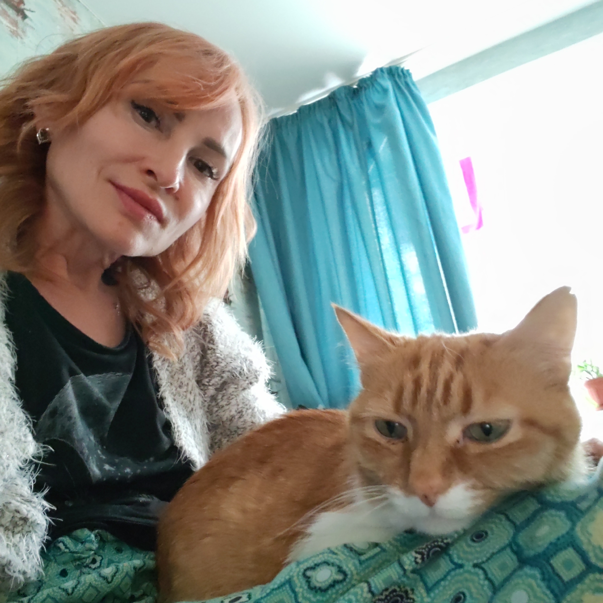 An indoor selfie captures a woman and an orange tabby cat in Chaikovsky, Russia. The woman, positioned at the top left, has reddish-blonde hair, light skin, and is wearing a dark top beneath a light, fuzzy cardigan. She has visible eyeliner and lipstick, and a small earring is visible on her left ear. Her expression is a gentle smile, and she is looking directly at the camera.
The orange tabby cat, with green eyes and white markings on its chest and muzzle, is resting comfortably in the foreground, seemingly on a patterned blanket or fabric that features teal, green, and dark blue geometric and circular designs. The cat appears calm and relaxed.
The background shows a brightly lit room, indicative of daytime. Light blue ruffled curtains are prominent behind the subjects, suggesting the presence of a window. A small, potted green plant is faintly visible in the far right background. The walls appear light-colored. The image conveys a serene and intimate moment between the woman and her pet. No visible text is present.