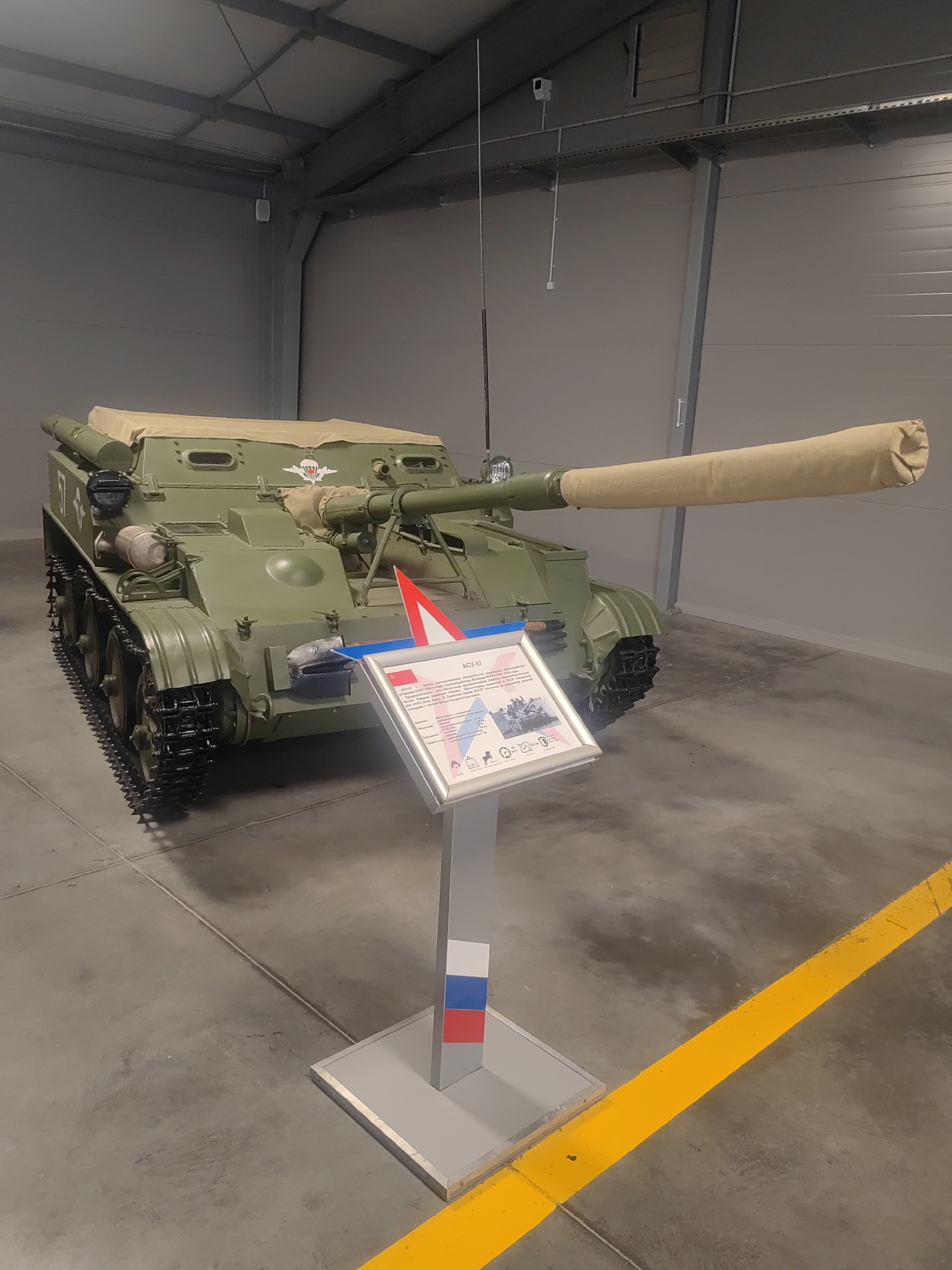 This image showcases a Soviet-era ASU-57 airborne assault gun, displayed indoors on a concrete floor. The vehicle is painted in a military green camouflage. It has a long gun barrel covered with a tan canvas sleeve and is equipped with tracks for mobility. A tarpaulin covers the top portion of the vehicle. In front of the ASU-57, a display stand is positioned, featuring information about the vehicle. The stand has a graphic design that includes what appears to be the Russian flag colors. The overall setting is a well-lit indoor exhibition space, likely a museum or exhibition hall, with industrial-style ceiling structures and gray walls. The lighting suggests it is daytime, and there are no external environmental cues like weather or natural light visible. The scene is static, as the vehicle is on display and not in operation. The context provided places this scene in Kubinka, Russia.
