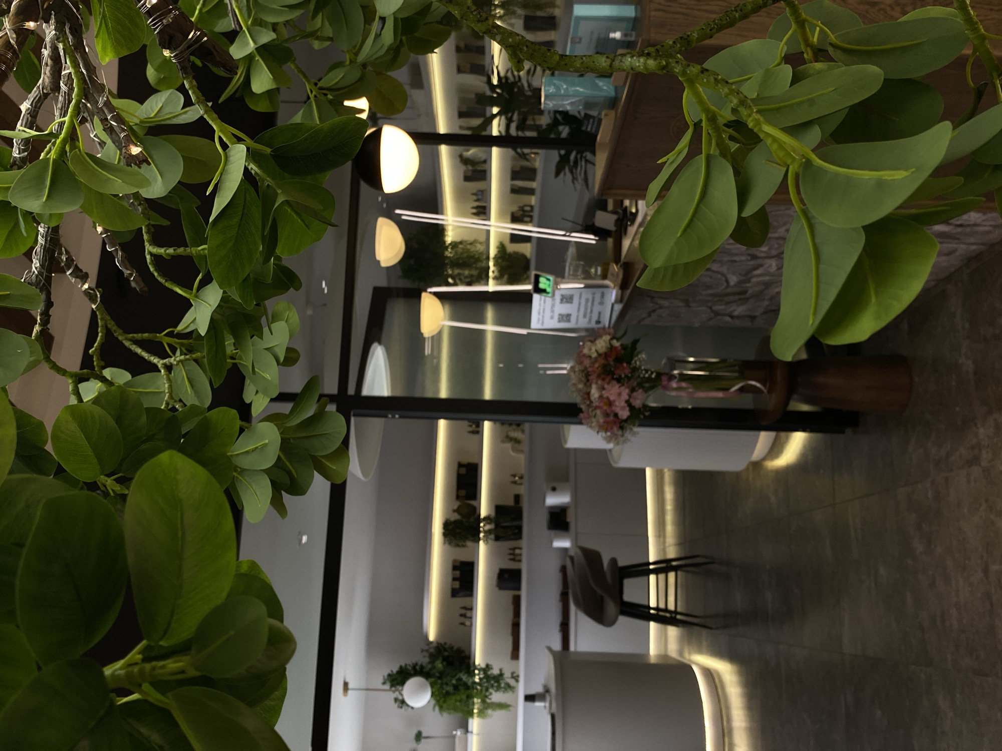 A brightly lit modern interior is partially obscured by foreground greenery. The space features white shelving lined with products, accent lighting, and potted plants. A counter is visible with a vase of flowers. The scene includes bar stools and a clean, minimalist aesthetic. Based on the style, this could be a commercial space, such as a boutique or a salon. The location in Votkinsk, Russia cannot be confirmed with this image alone.