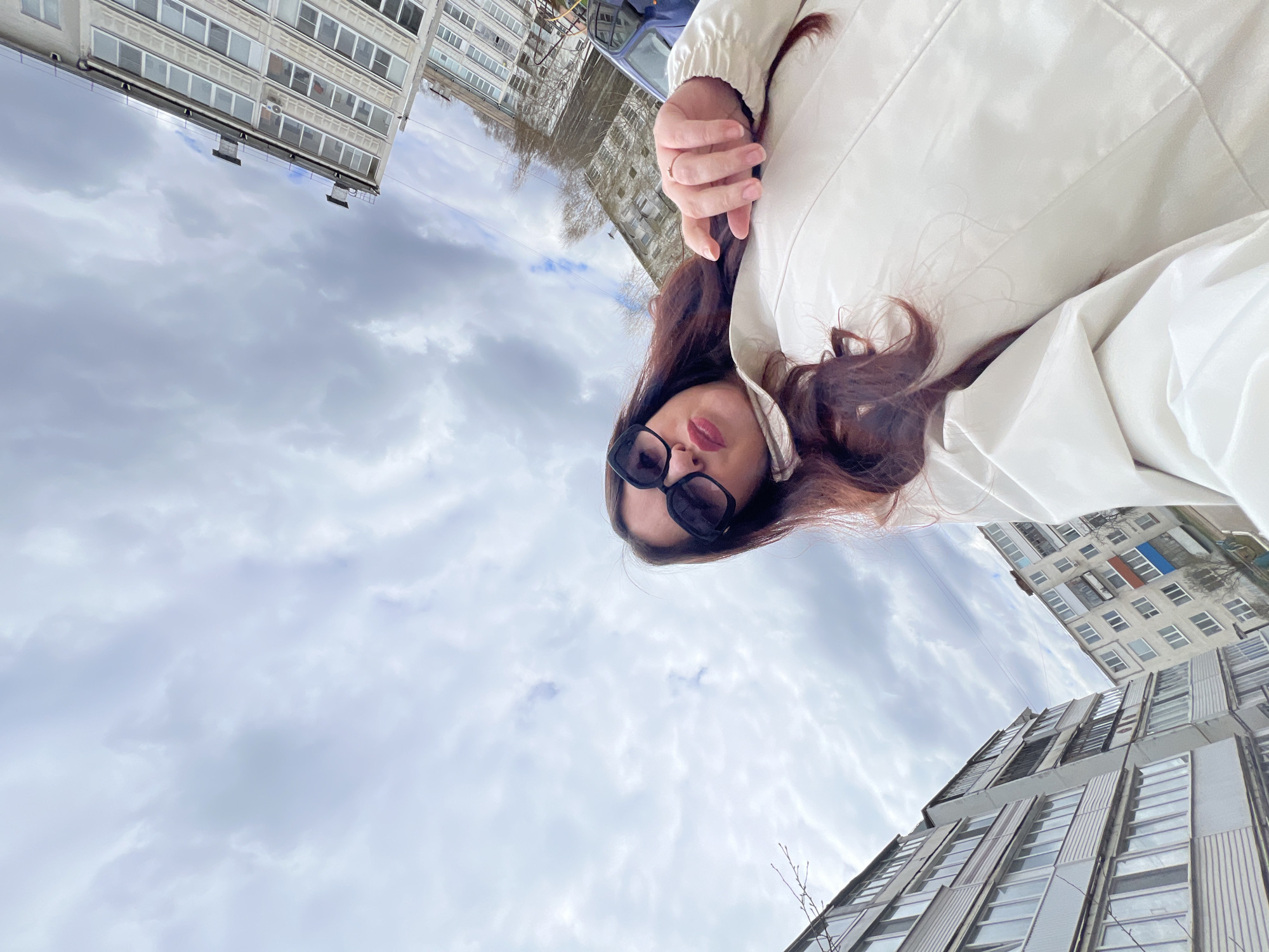 A young woman is depicted outdoors in an urban setting, likely Noviy, Russia, based on the provided context. The image is taken from a low angle, looking upwards. The woman, with long, dark brown hair, is wearing black sunglasses and a light-colored (possibly white or cream) jacket. Her left hand, adorned with a ring on the ring finger, is visible near her chest. Her lips are slightly pursed as she looks directly towards the camera.
The background features several multi-story residential buildings with numerous windows and balconies, typical of apartment blocks found in post-Soviet architecture. The buildings appear to be made of concrete or light-colored masonry, with some showing signs of wear or discoloration. One building in the mid-ground has an air conditioning unit visible on a balcony. The sky above is overcast, filled with soft, diffused clouds, suggesting a cloudy daytime. There is no visible text or identifiable logos in the image. The overall impression is a candid shot in an everyday urban environment.