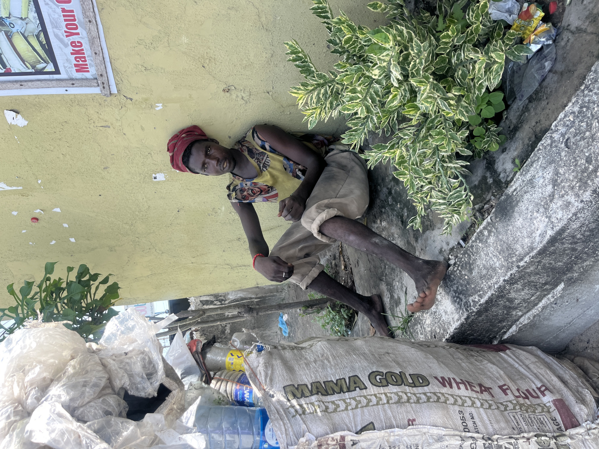 In Ado, Nigeria, a person with dark skin sits on a concrete ledge against a yellow wall. The person wears a red head covering, a patterned tank top, and rolled-up beige pants, with bare feet. Their left hand rests on their head, and they are looking towards the viewer. Next to them, greenery grows. Below the ledge, a pile of plastic bags and water bottles sits against a "Mama Gold Wheat Flour" sack. A sign with the phrase "Make Your" can be seen at the top left.