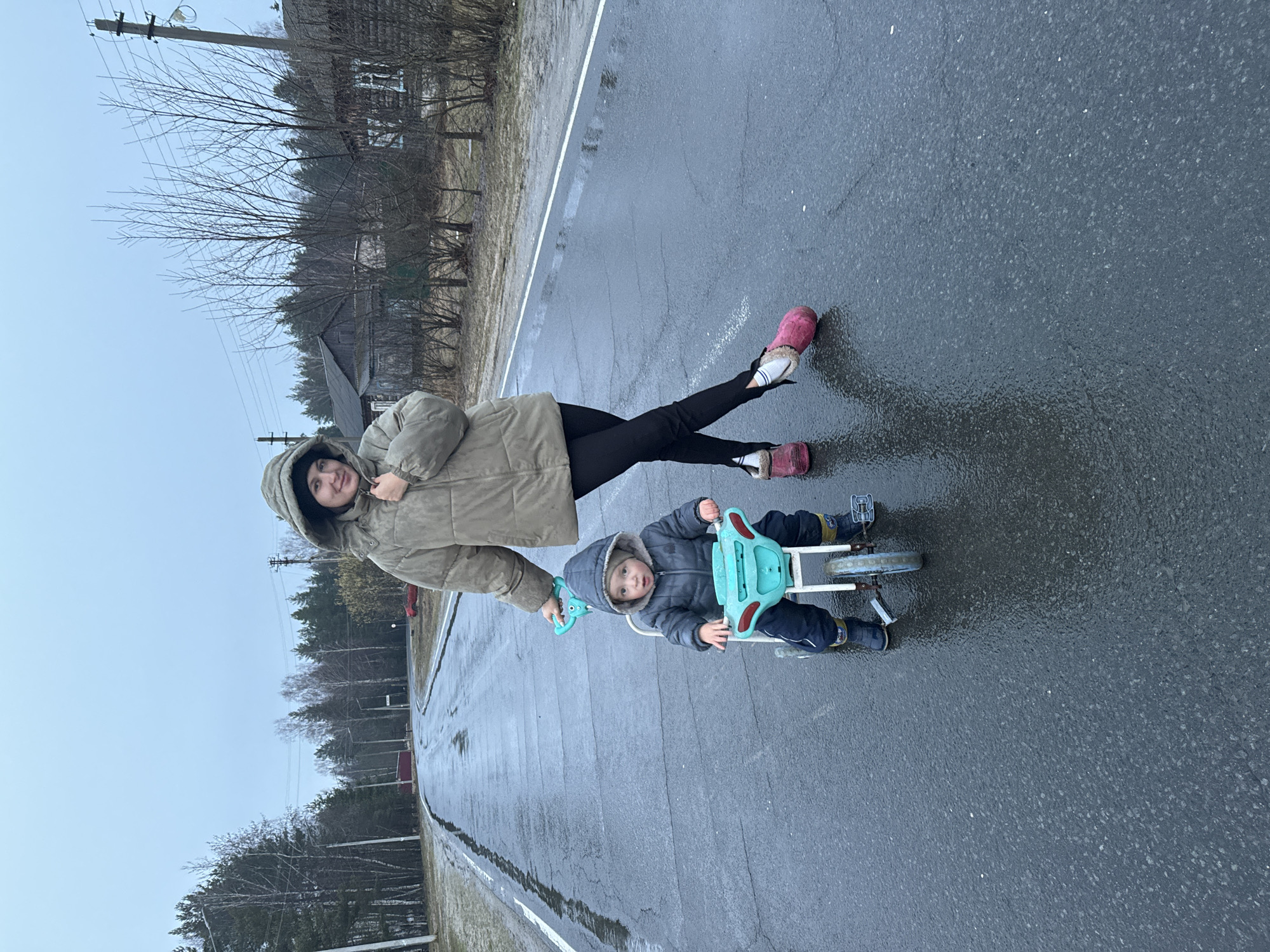 A woman and a young child are on a wet road in what appears to be a suburban area. The woman, wearing a hooded jacket and black pants, is holding the handle of a blue tricycle. The child is sitting on the tricycle, wearing a hooded jacket. The road is gray and wet, reflecting the sky, and is marked with a white line. Trees and buildings are visible in the background, suggesting a residential neighborhood. The weather appears overcast.