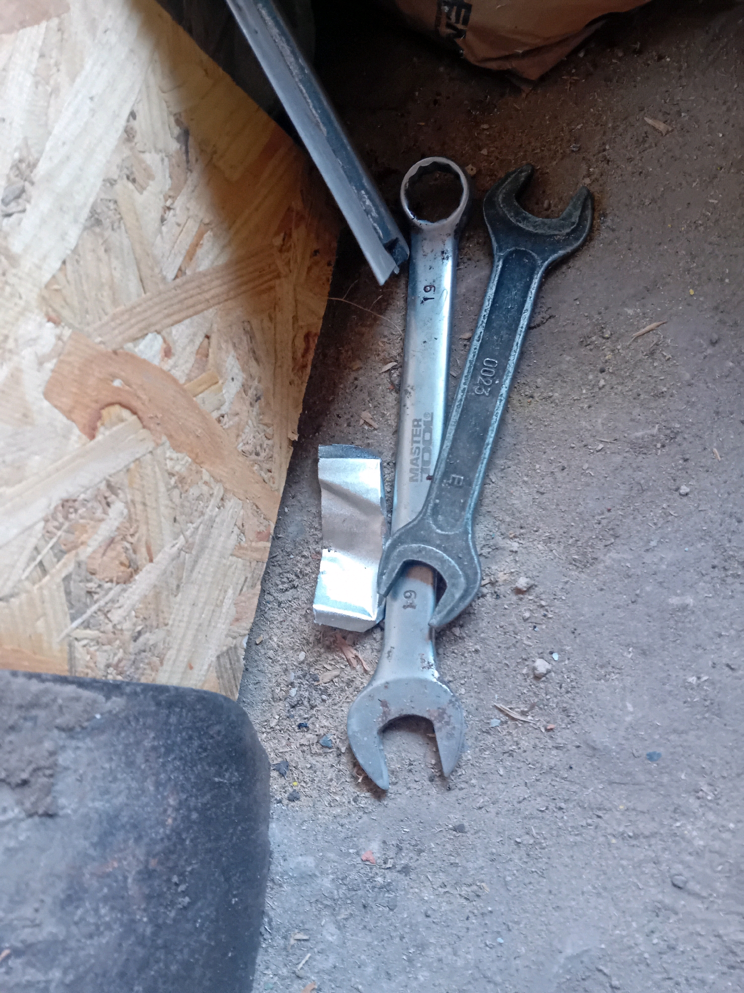 A pair of open-ended wrenches and a piece of metallic foil are on a dirt ground next to a piece of oriented strand board. The wrenches are positioned diagonally, one on top of the other, with their open ends pointing towards the bottom right of the frame. The top wrench is silver and has the number "19" stamped on it. The bottom wrench is darker, possibly black or dark gray, and has the markings "MASTER TOOL" and "0023" and "19". The foil is a crumpled, silver rectangular piece situated between the wrenches. In the background, a portion of a brown paper bag is visible, along with the edge of a dark, possibly black, cylindrical object, which might be a tire. The scene is lit from above, casting slight shadows. This image does not provide any specific information or context about the city of Chuhuiv, Ukraine, or any details about people, actions, or interactions.
