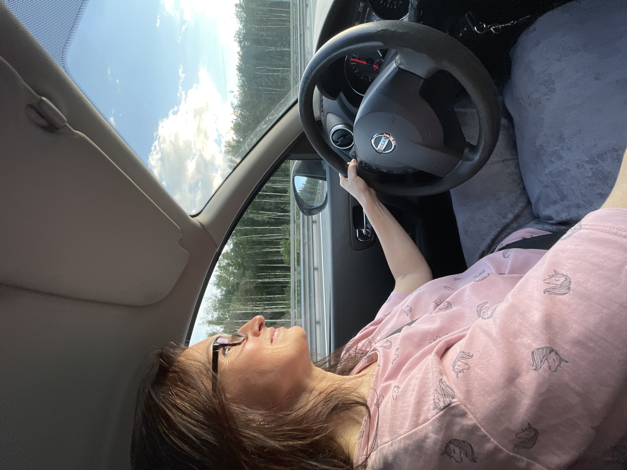 A woman with brown hair and glasses is driving a car. She is wearing a pink t-shirt with a unicorn print and grey pants. The car is on a highway with a forest on the right side and a blue sky with clouds above. The steering wheel has a Nissan logo. The dashboard shows the time as 20:40. The image appears to have been taken during daylight hours, likely late afternoon or early evening, given the lighting and time displayed on the dashboard. The location is identified as a highway in Saint Petersburg, Russia.