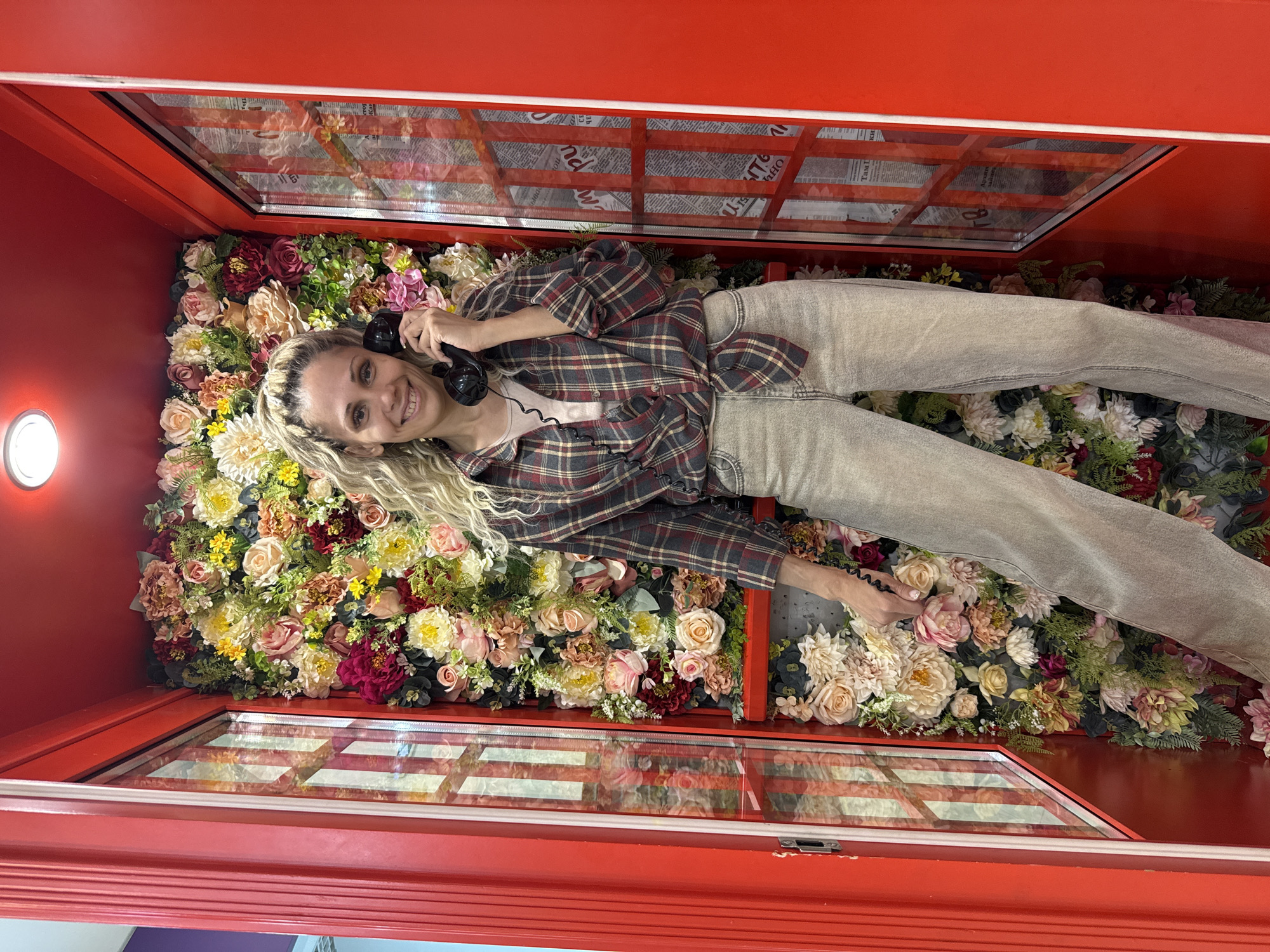 A woman lies inside a red phone booth decorated with flowers. She is holding a black telephone receiver to her ear and smiling at the camera. She wears light-colored jeans and a maroon and grey plaid shirt tied at the waist over a pink top. The phone booth has windows with newspaper clippings behind the glass. A light fixture is visible in the top left corner of the booth. The phone booth is located in Шебунино, Russia.