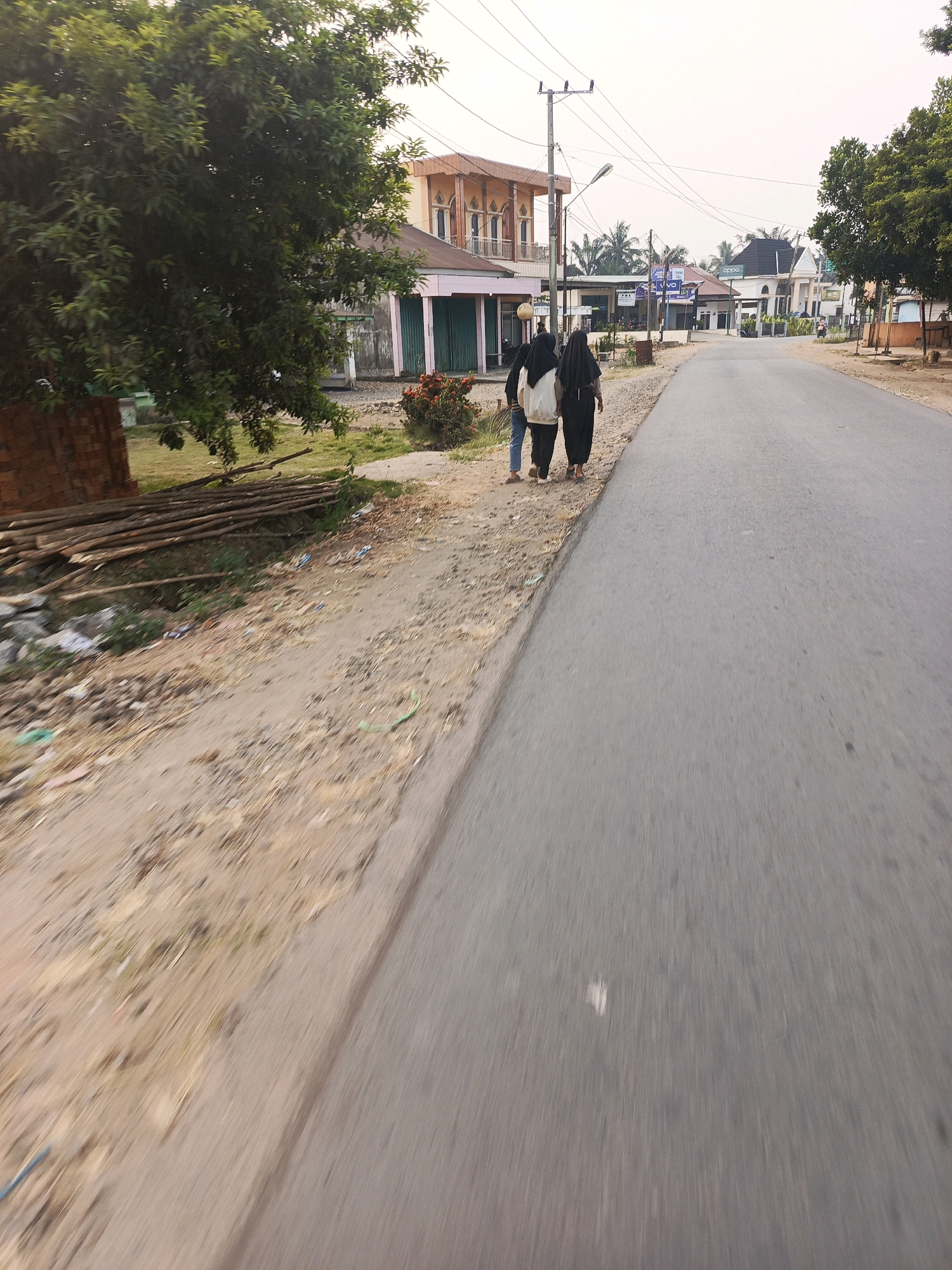 Two individuals, likely women based on their attire, walk away from the camera on the left side of a paved road. They are both wearing dark-colored headscarves and loose-fitting clothing. The person on the left wears a white jacket over dark pants, while the person on the right wears all dark clothing. They are walking on a dirt shoulder next to the road. The environment appears to be a rural or semi-urban area in Megang Sakti I, Indonesia. On the left side of the road, there are buildings with businesses, including one with green roll-up doors and another multi-story building with balconies and arched windows. There are also trees, a stack of logs, and some scattered debris on the ground. Electrical poles and wires are visible overhead. The road itself is asphalt and appears to be in moderate condition. The sky is overcast and the overall lighting suggests it is either early morning or late afternoon. The perspective of the image indicates it was taken from a moving vehicle or by someone walking behind the subjects.