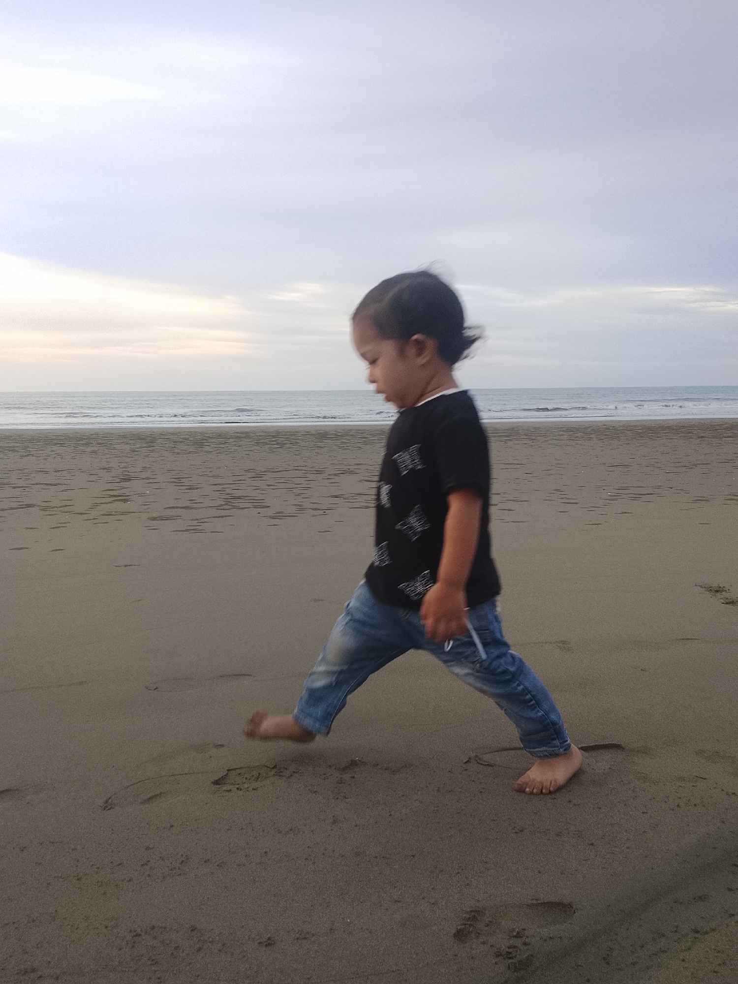 A young child walks on a sandy beach, their left foot leaving a visible footprint in the sand. The child wears a black t-shirt with a white pattern and blue jeans. The ocean is visible in the background with gentle waves rolling towards the shore under a cloudy sky. The scene appears to be set during the late afternoon or early evening, given the soft lighting. The location is identified as Reudeuep, Indonesia.