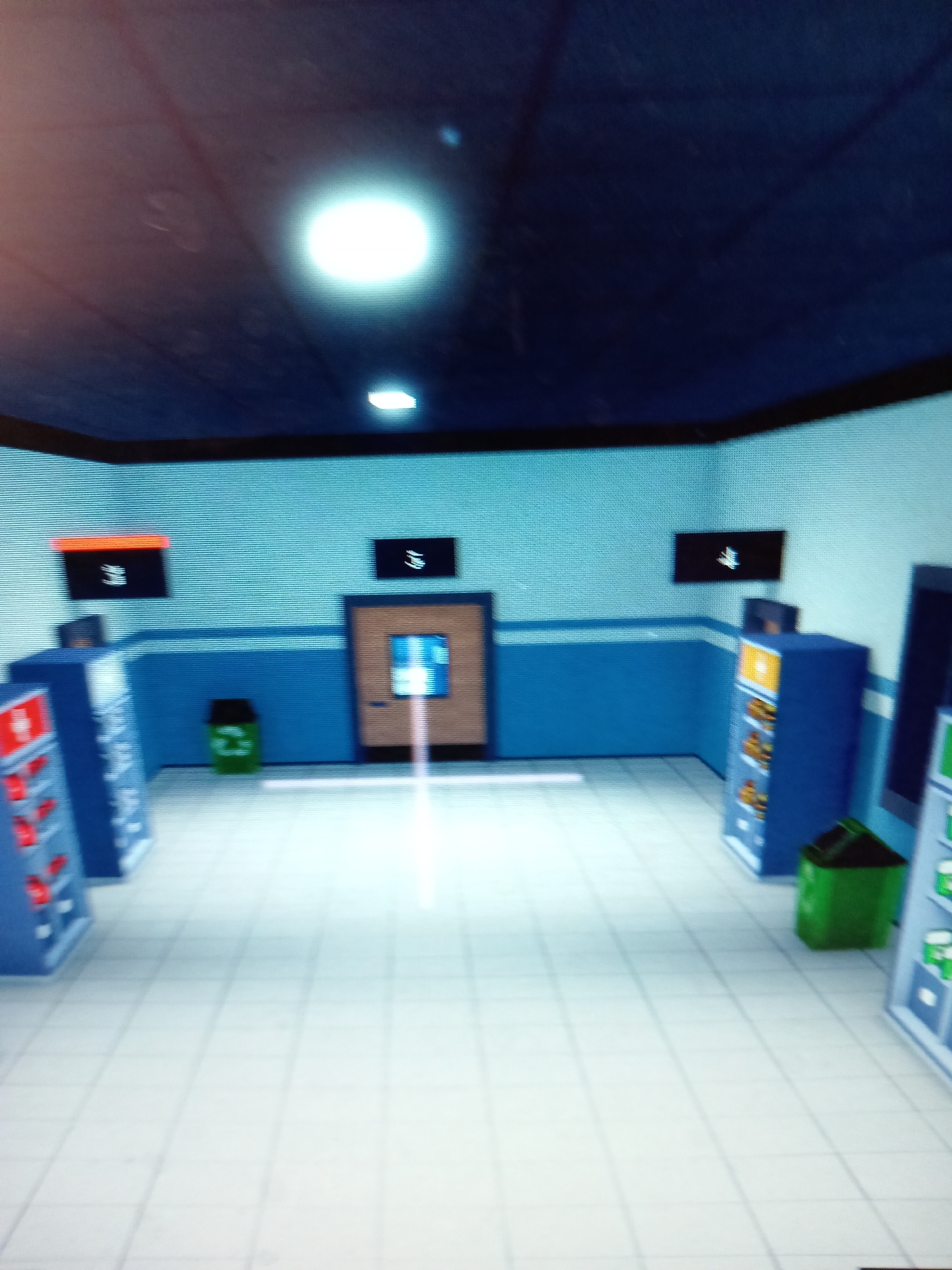 The image depicts an indoor scene, possibly a hallway or common area, with bright fluorescent lighting from the ceiling. The walls are painted in alternating bands of light blue and white. To the left, there are two blue display units filled with items, one with red compartments. In the center, there is a wooden door with a window, and in front of it, a light beam extends onto the tiled floor. On the right, there are two more blue display units, one containing items in yellow and orange hues, and a green trash bin with a recycling symbol. Several black rectangular screens are mounted on the walls at various heights, displaying white symbols or text. The floor is tiled with a grid pattern in a light color. The overall impression is of a clean, modern, and somewhat sterile environment. There are no visible people or animals in the scene. The location in the city of Rudny, Kazakhstan, is not discernible from the visual elements present in the image.