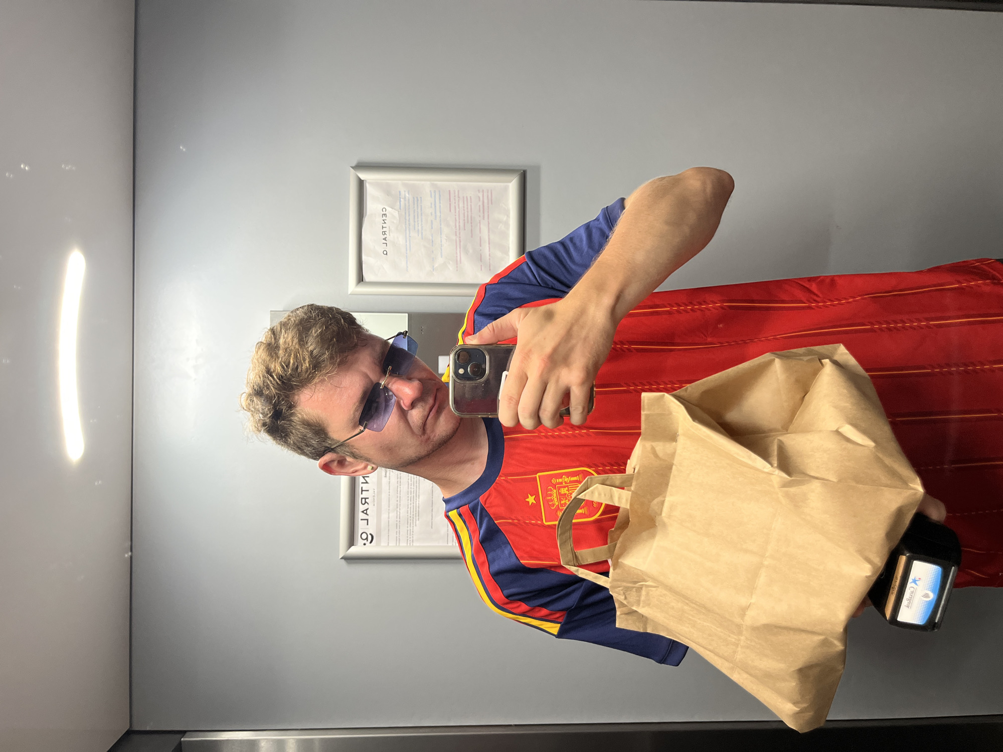 A man is taking a mirror selfie in an elevator. He is wearing a red and blue Spanish national football team jersey and blue-tinted sunglasses. He is holding a brown paper bag in his left arm and a smartphone in his right hand, which he is using to take the photo. The elevator has gray walls and a brightly lit ceiling. There are framed documents on the wall behind him. A contactless payment terminal is visible underneath the paper bag. The overall scene appears to be in an urban environment, possibly a public building or residential complex in Quart de Poblet, Spain.