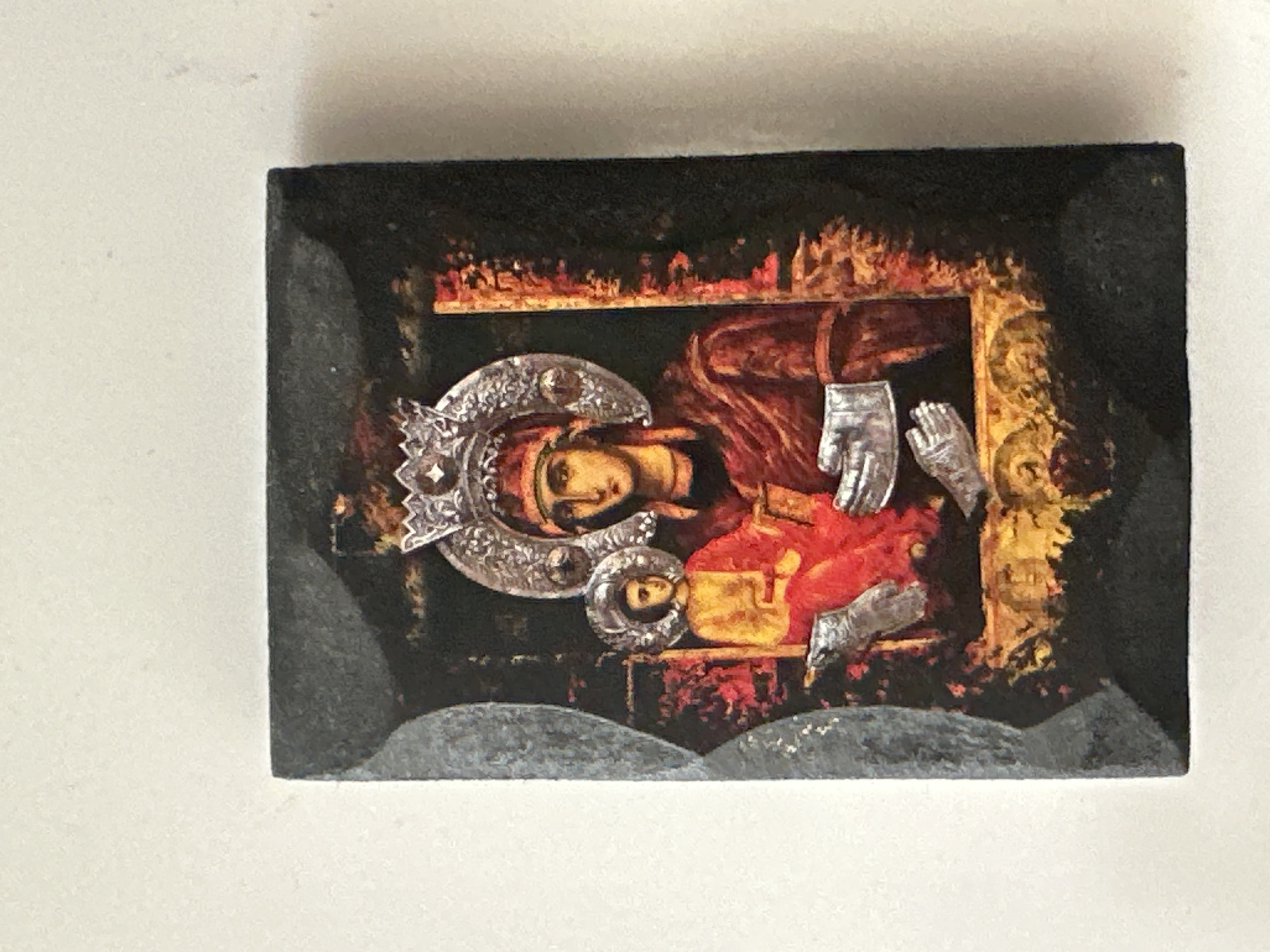 The image shows a religious icon, likely of the Virgin Mary and the Christ child, housed in a dark, ornate frame. The icon itself is small and rectangular, with a central depiction of two figures. The figure on the right, presumably the Virgin Mary, wears a red garment and a crown. The figure on the left, the Christ child, is dressed in yellow and red. Both figures have halos. The frame surrounding the icon is dark gray and appears to have a textured or marbled finish. The icon is placed on a light-colored, textured surface, possibly a wall or table. The lighting is bright and comes from the front, casting subtle shadows. There are no identifiable people or landmarks in the scene, and the location is unspecified.