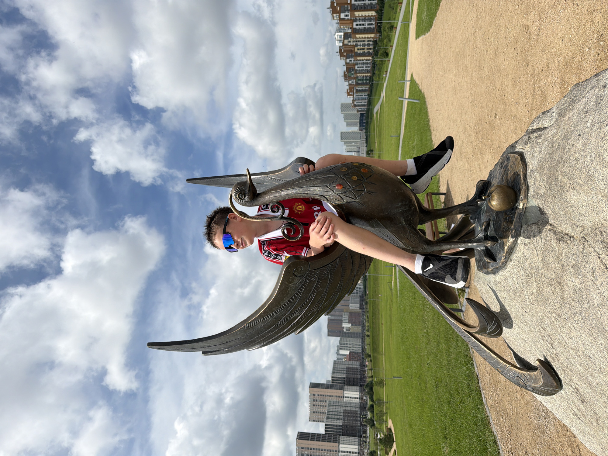 A young boy with short brown hair and blue sunglasses is sitting on a bronze sculpture of a bird-like creature with large wings. He is wearing a red and white football jersey with a crest on the left side of his chest and headphones around his neck. His legs are bent, and his feet are resting on the sculpture. The sculpture is perched on a large, rough-hewn stone. In the background, there is a grassy park with a path and several tall, modern apartment buildings. The sky is blue with scattered white clouds. This is an outdoor scene in a park in the city of Yekaterinburg, Russia, likely in the Zalesye residential complex, given the distinctive architecture of the buildings and the presence of this sculpture.