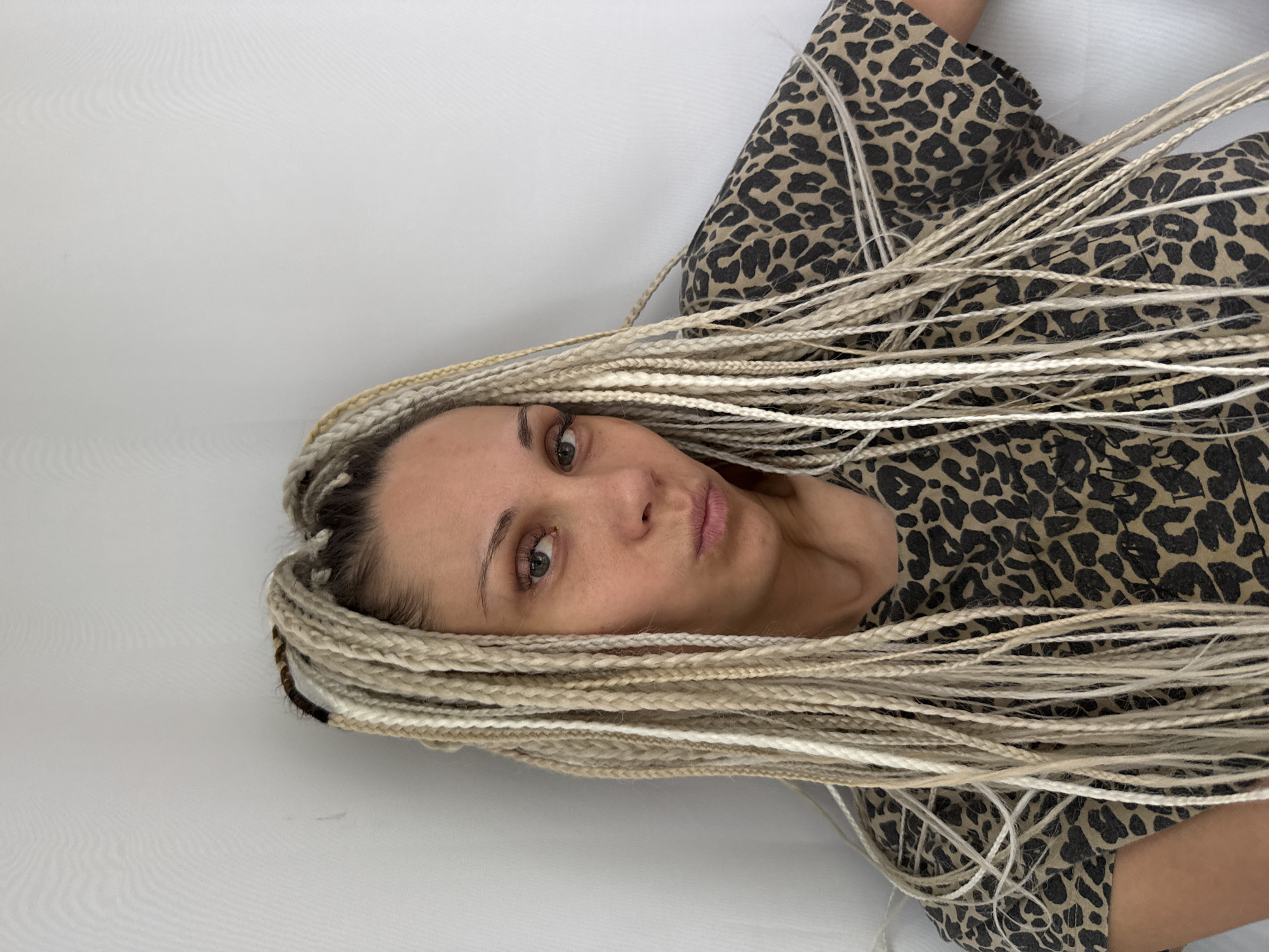 A woman with blonde box braids is posing for a photo against a white wall. She is wearing a leopard print t-shirt. The woman is looking directly at the camera with her lips slightly pursed. Her head is tilted to her right. The image is a medium shot, focusing on her face and upper body. There is no discernible location in the city of Rusanovo, Russia visible in this image.