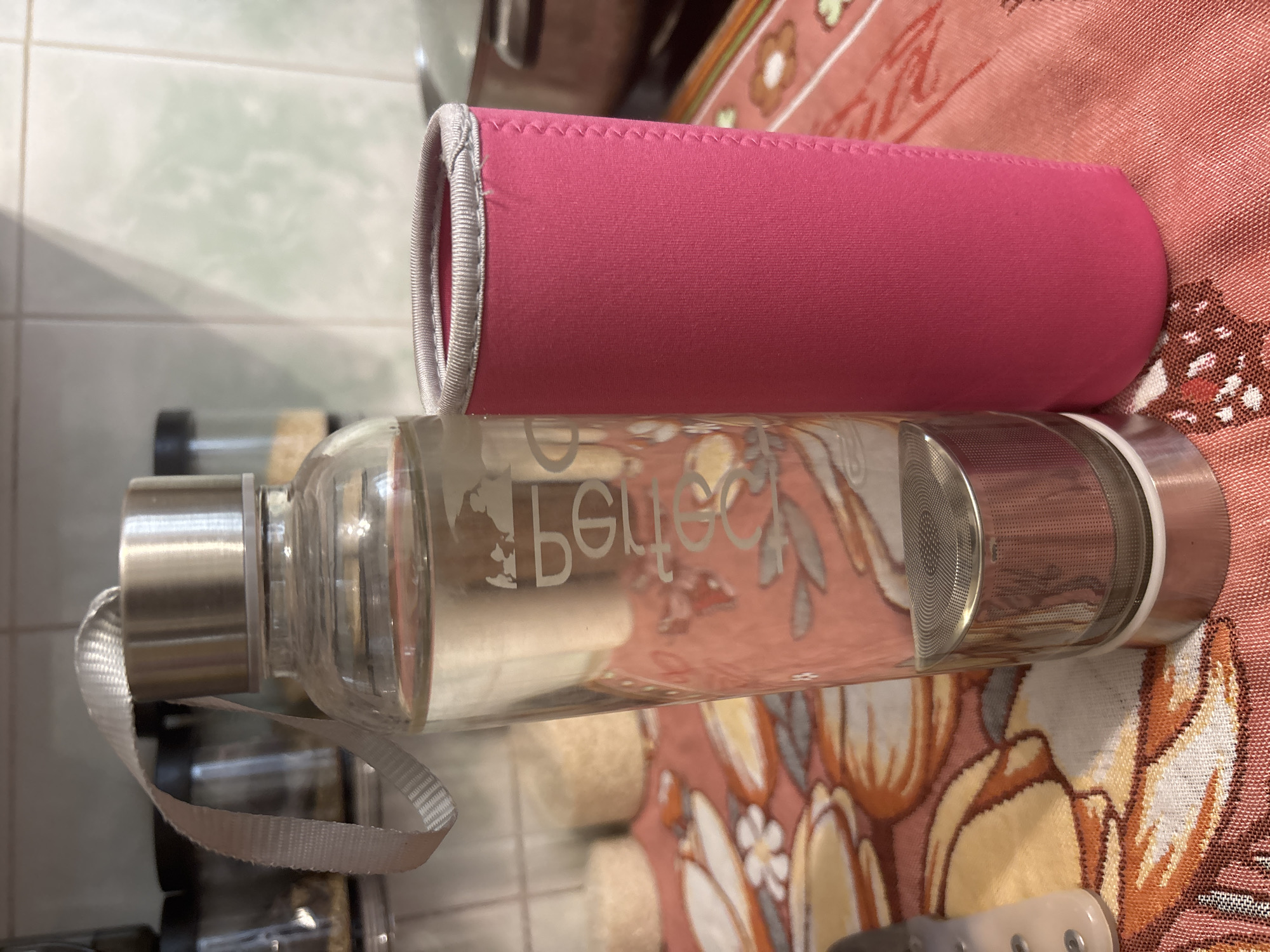 A glass water bottle is lying horizontally on a surface covered with a floral-patterned fabric. The bottle has a silver-colored metal cap with an attached white fabric loop. A pink cylindrical sleeve is partially covering the middle section of the bottle. The words "bebetter" are faintly printed on the bottle's surface. The background shows a tiled wall and other bottles, suggesting the scene is indoors. There is no indication of the image's location being Zagatuy, Russia.