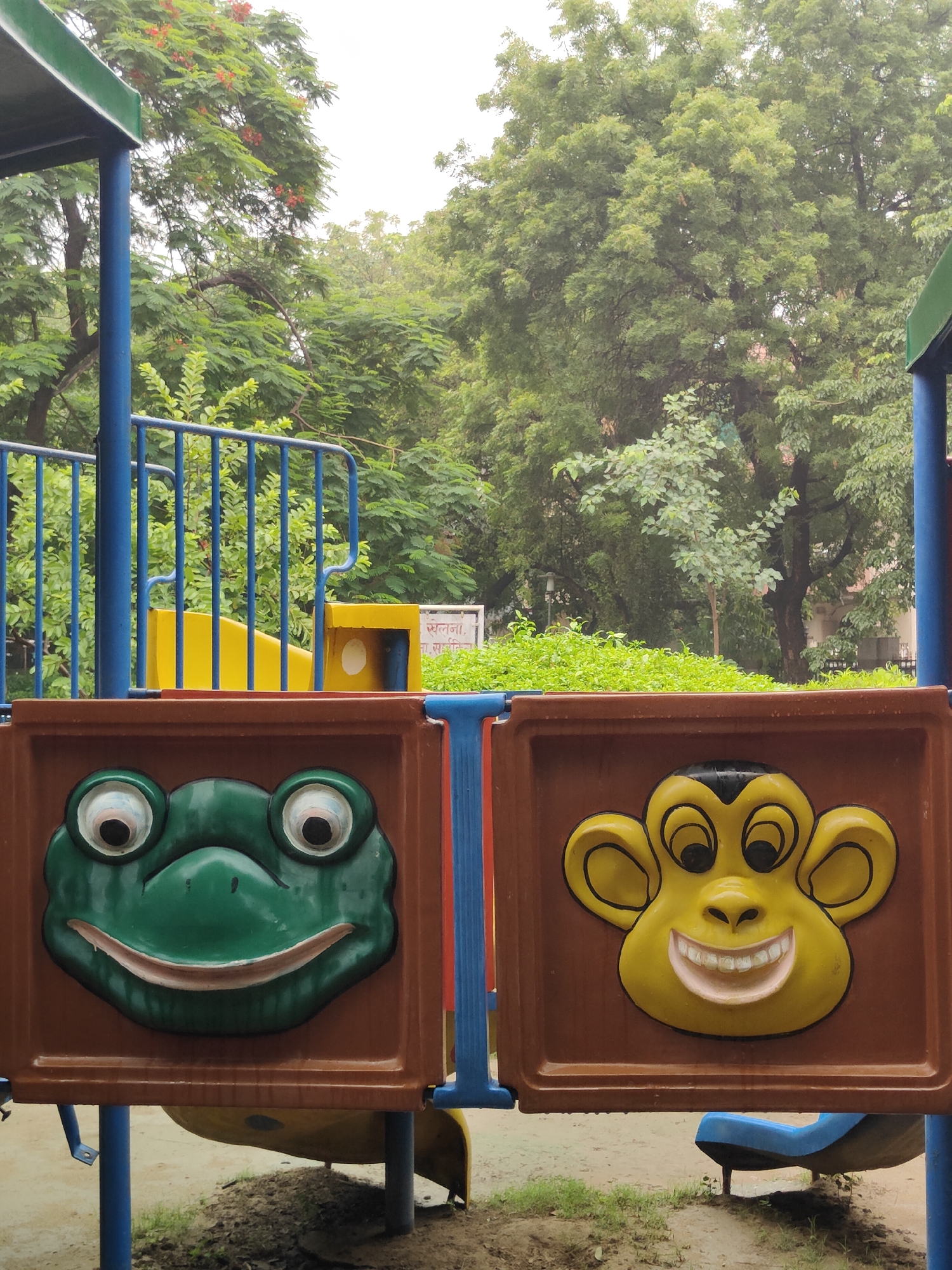 The image depicts a close-up view of a playground structure at a government school in Gurgaon, India. The playground features colorful panels with molded faces of a frog and a monkey. The frog panel is green with large, bulging eyes and a wide smile. The monkey panel is yellow with prominent ears, black eyes, and a wide, toothy grin. The panels are framed in brown and blue. Behind the playground structure, a yellow slide is visible, along with blue railings. In the background, lush green trees and foliage dominate the scene, suggesting an outdoor environment. A white sign with Hindi text is also partially visible behind the playground, indicating a park or recreational area. The ground appears to be a mixture of dirt and grass.
