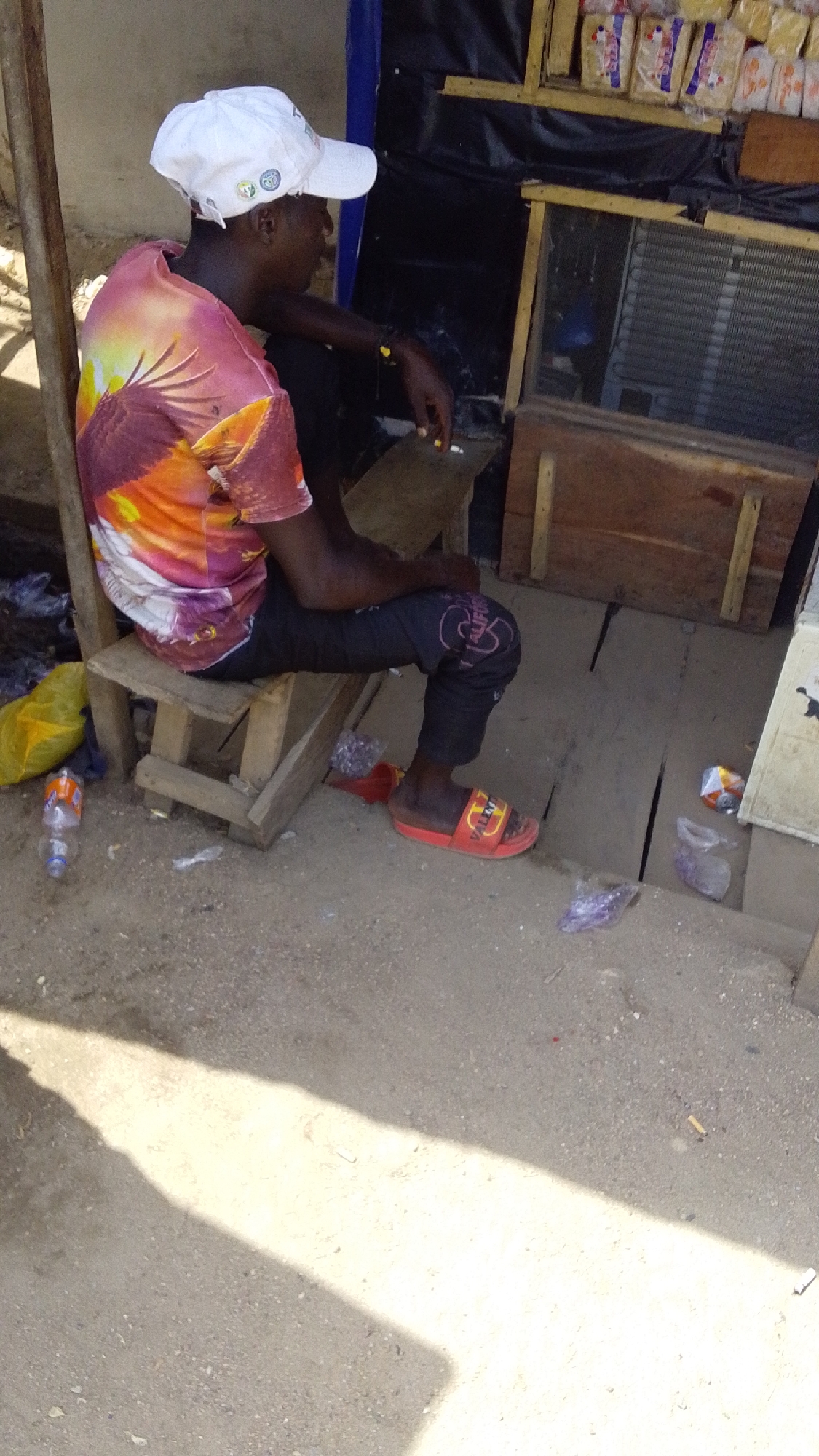 The image depicts a man sitting on a small wooden stool in Lokoja, Nigeria. He wears a white baseball cap, a t-shirt with a bird print in shades of red, orange, and yellow, black pants with designs on the knee, and red slides. He holds a cigarette in his hand. The man is situated next to a wooden stall or shop. Behind him are various items including packaged bread and a windowed refrigeration unit. A yellow plastic bag and a water bottle are on the ground near the stool. Litter, including plastic wrappers and cigarette butts, is scattered around the area. The ground is a mixture of concrete and dirt, with a patch of sunlight shining on it.