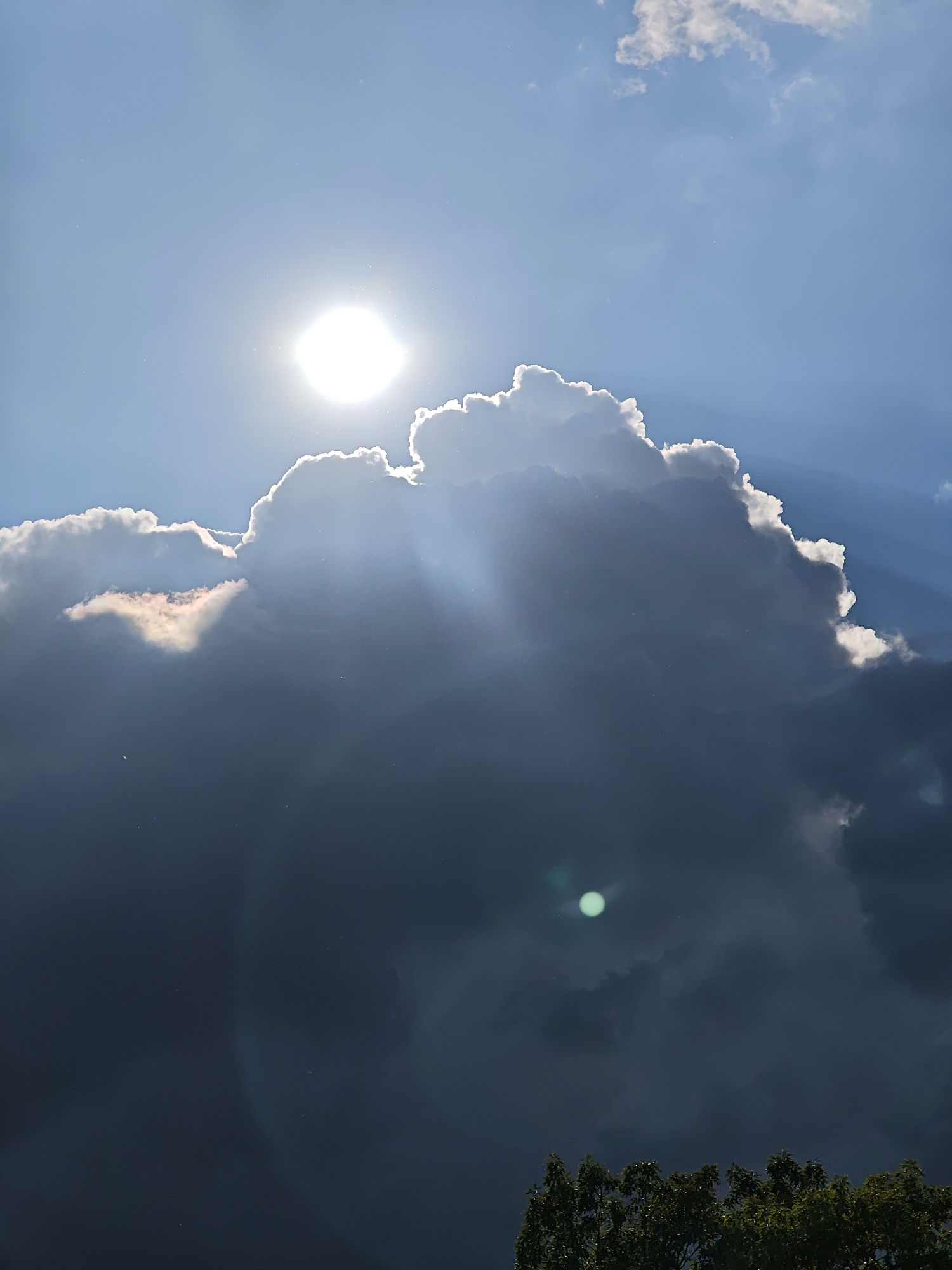 The image depicts a daytime scene with a bright sun partially obscured by a large, dark cumulonimbus cloud. Light rays are visible emanating from behind the cloud. The sky is blue in areas not covered by the cloud. In the bottom part of the image, the tops of trees are visible. There is no indication of location in Vratsa, Bulgaria, nor are people or interactions depicted in the image.