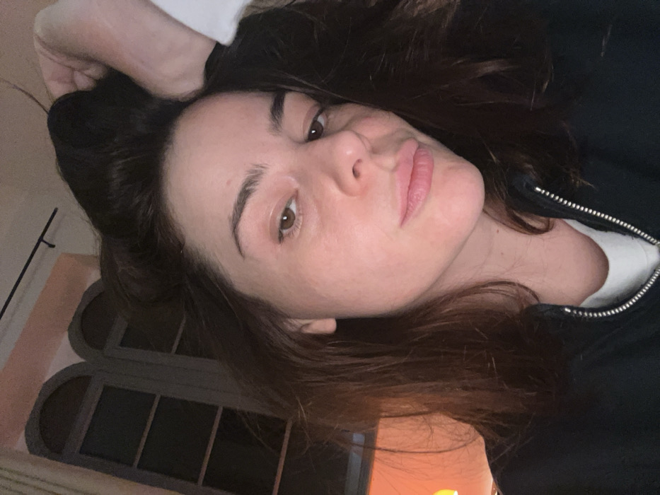 A woman with brown hair and fair skin looks toward the camera. She has brown eyes, arched eyebrows, and full lips. She wears a dark zippered jacket over a white t-shirt. Her hair is slightly disheveled. The background includes a window with a dark interior and architectural details. The photo is taken at a tilted angle, creating a casual or candid feel.