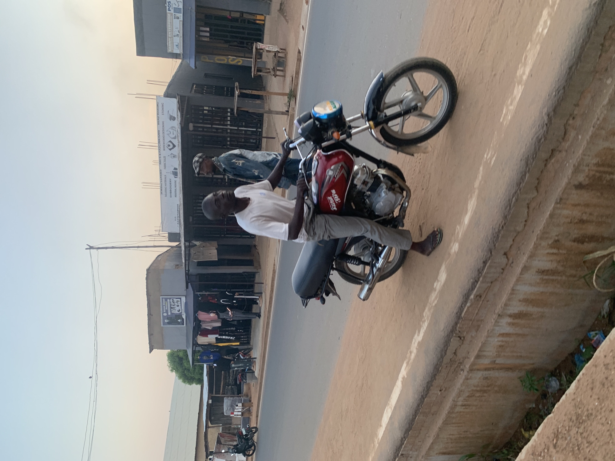 The image depicts a man seated on a red motorcycle parked on a roadside in Sabojibi, Nigeria. The man is wearing a white shirt and light-colored pants. Behind him, another man stands on the street. On the left side of the road, there are small shops and businesses. A sign for "SAMMY ENTERPRISE POS" and "WINNERZ POS" is visible on one building. There is a sign for "Artess Klassic" describing sewing classes and services like bead making, wireworks designing, fabric sales, embroidery, and industrial weaving. Several other motorcycles are parked along the road in the background. The environment appears to be a commercial area. The lighting suggests it's daytime, possibly late afternoon.