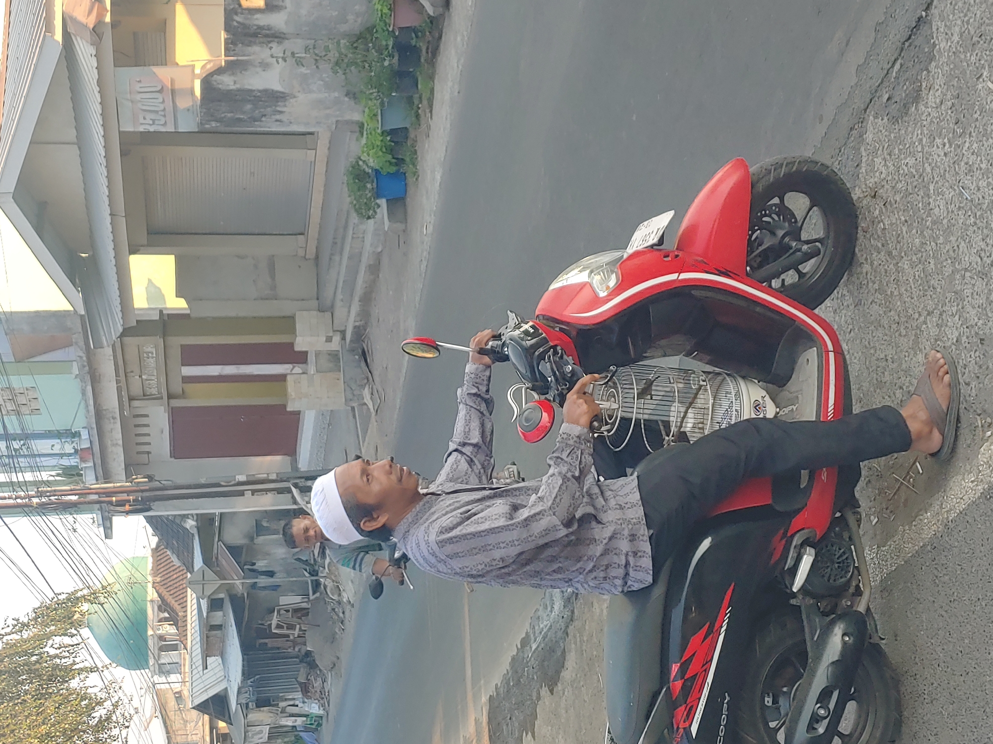 In Kembang Kerang, Indonesia, a man in a white cap and a patterned gray shirt is riding a red and black scooter. The scooter has a cage attached to its front, and the man is wearing black pants and brown sandals. He is looking to his right, with his hands on the handlebars. Behind him, another man on a scooter is visible. The scene takes place on a paved road lined with buildings and trees. The sun is casting a warm light on the buildings, and power lines are strung across the sky.