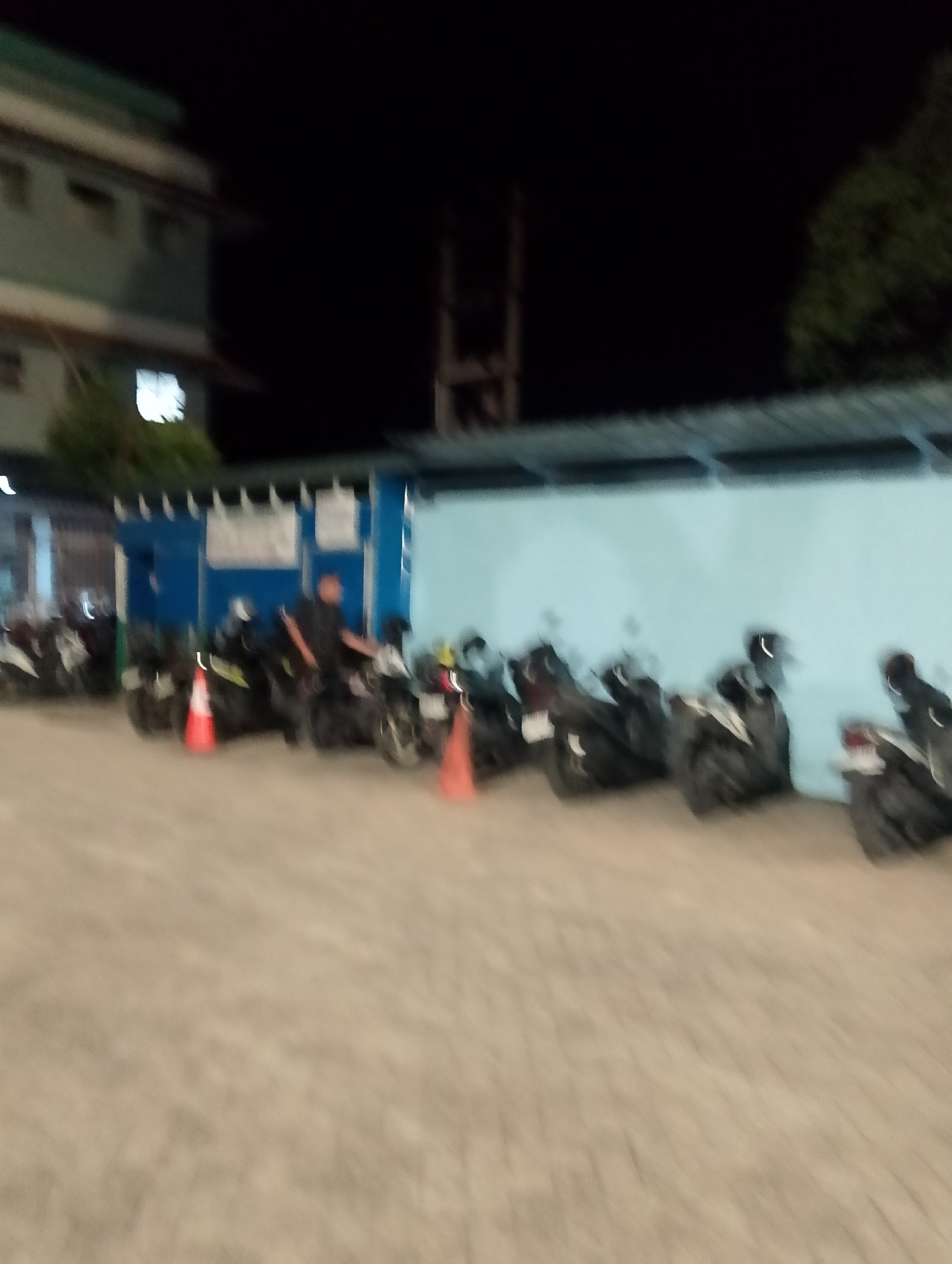 This is a blurred, night-time photograph of a row of parked motorcycles in what appears to be an outdoor parking lot. The background includes a light blue wall, a building with a green and white facade, and trees. There is a person standing next to one of the motorcycles, facing away from the camera. Two orange traffic cones are visible in the foreground, placed between some of the motorcycles. The image is likely from the City of Pematang Siantar, Indonesia, given the provided context.