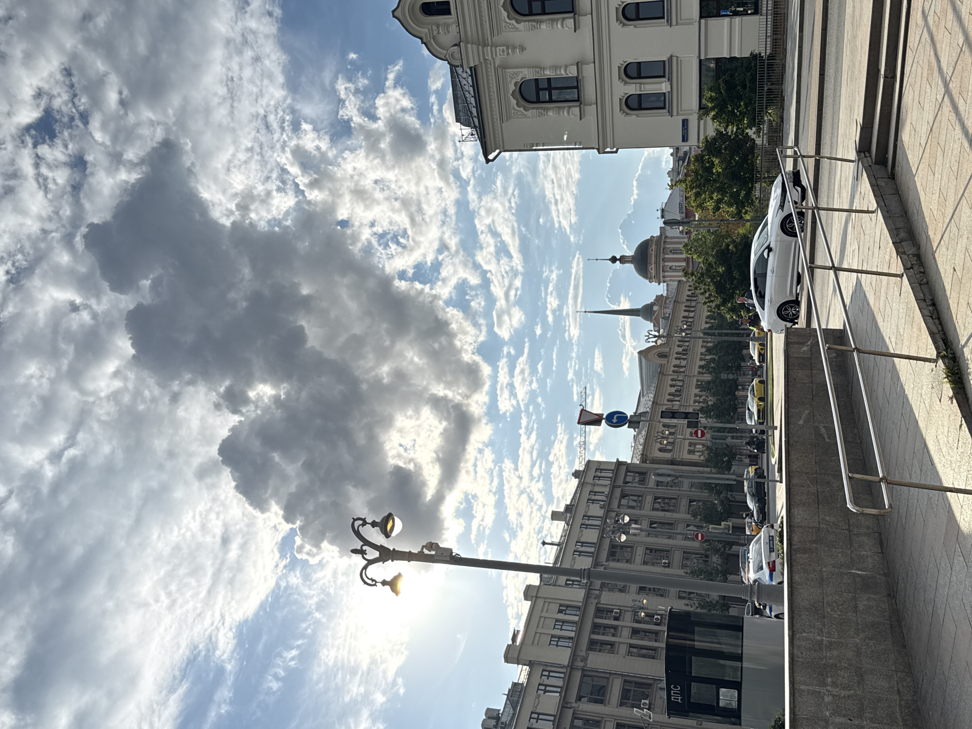 The image captures a street scene in Moscow, Russia, under a cloudy sky. The orientation of the image is vertical, with the sky occupying the majority of the frame and the cityscape below. Large, dark clouds dominate the upper left portion of the sky, contrasting with patches of bright blue and scattered white clouds elsewhere. Sunlight filters through the clouds, creating a dramatic effect.
Several buildings are visible, showcasing architectural styles typical of urban environments. In the foreground, a sidewalk with a metal railing leads up a set of stairs. Adjacent to the stairs, a white car is parked. Further down the street, more buildings with multiple windows line the road, and trees provide greenery. A streetlamp with two lights stands prominently in the mid-ground, illuminated despite the cloudy conditions. Traffic lights and road signs, including a blue circular sign with a white arrow, are also visible.
Several cars, including a white one that appears to be a police vehicle, are on the street. Some people can be seen near the white car. The overall impression is a dynamic urban environment on a day with mixed weather. The time of day appears to be late afternoon, given the angle of the sunlight and the shadows cast. No specific emotions are conveyed, but the scene suggests a typical day in a bustling city. Visible text includes "ДПС" on a booth-like structure in the lower part of the image.