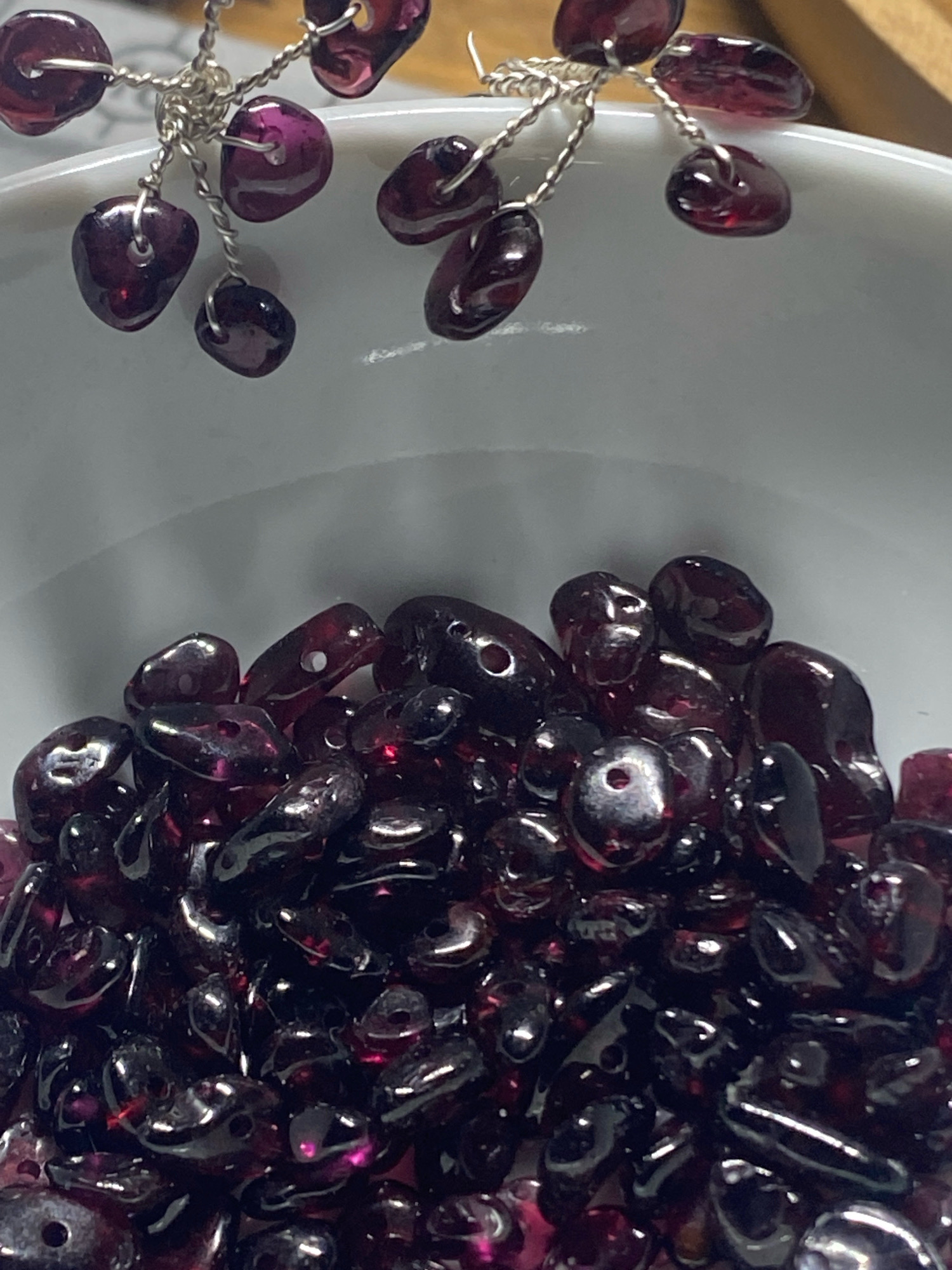 The image shows a close-up of a collection of dark red, translucent gemstones, possibly garnets, piled in a white bowl. Some of the gemstones are attached to a delicate silver wire structure at the top of the frame, suggesting they are part of jewelry or a decorative piece. The gemstones have irregular shapes and appear to be faceted or polished, reflecting light. The background is out of focus, with the white of the bowl being the most prominent element. There is no indication of a specific location in Kushchovskaya, Russia, or any contextual environmental details that would place it there. The scene is static and focuses solely on the gemstones.