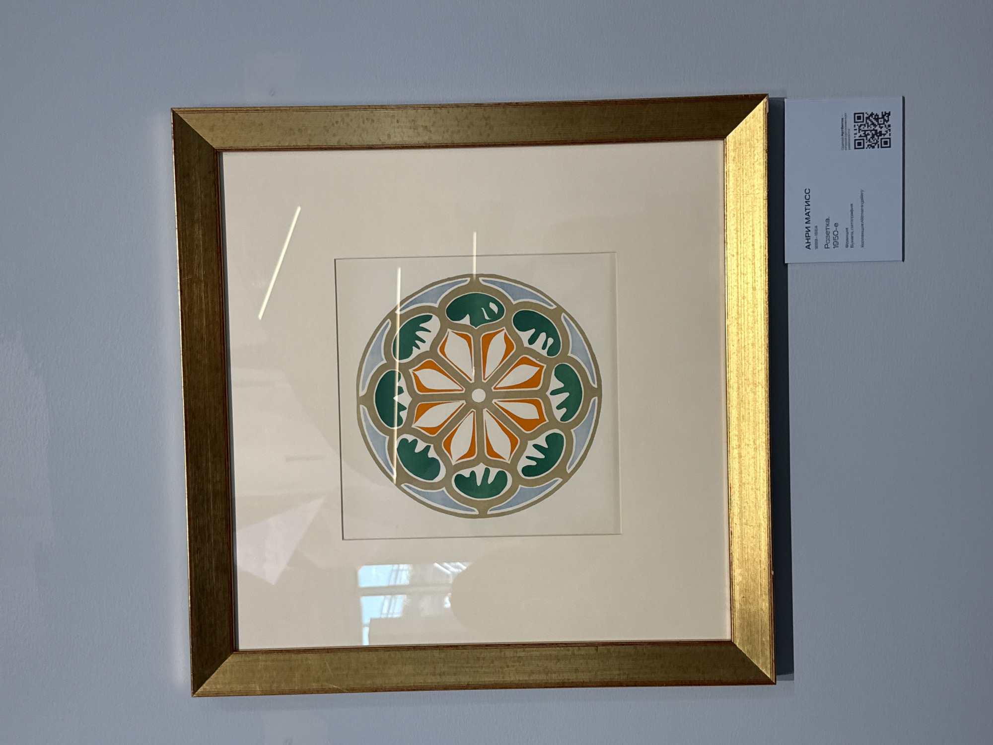 A framed artwork is displayed on a light blue wall. The artwork is a circular, abstract design with alternating orange and green petal-like shapes arranged around a central white circle. The design is framed by a light beige matting and a gold-toned frame. Below the artwork, a white label provides information about the piece. The label reads: "АНРИ МАТИСС 1869-1954 Ропетка. 1950-е Фрввция Бумага, литография Коллекция<Altmans gallery>". A QR code and additional text in Russian are also present on the label. The overall scene suggests an art exhibition.