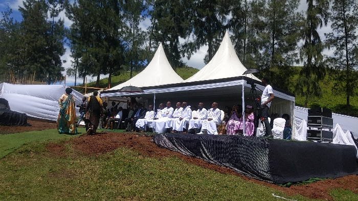 The image shows a scene in Rukoko, Democratic Republic of Congo (latitude: -1.624, longitude: 29.258). There are two white tents with black accents set up on a grassy slope. Under the tents, a group of men in white robes are seated in chairs, facing the camera. There is a woman in a pink dress seated to their right. Behind them, a man in a white shirt and black pants is holding a camera. In front of the tents, there is a black tarpaulin covered stage with a microphone stand and sound equipment. Two people in traditional attire are walking away from the tents on the left, and a few other people are seated on chairs to the right of the tents. The scene is outdoors, in a grassy area, surrounded by pine trees. The sky is partly cloudy.