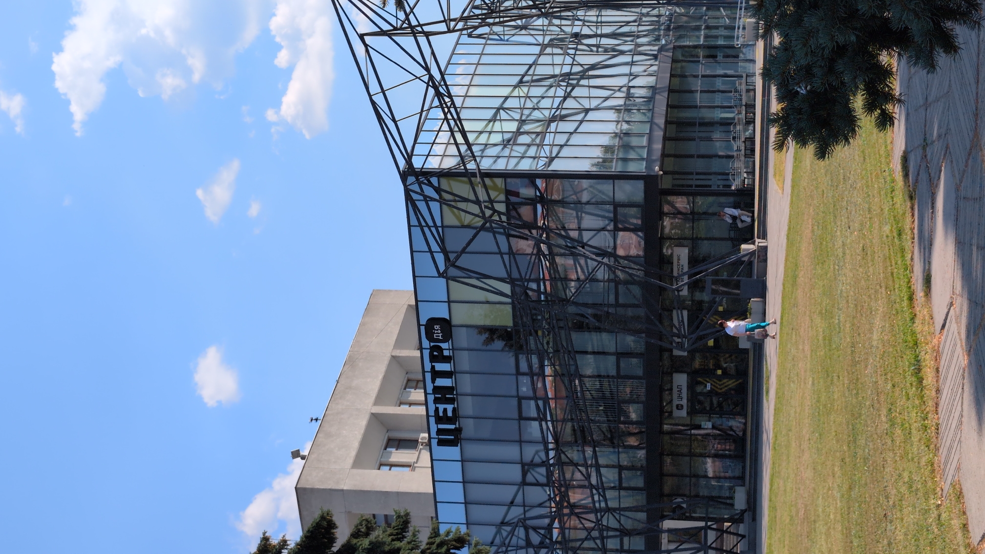 The image shows the exterior of a modern glass building in Poltava, Ukraine. The building features a prominent black metal geometric structure in front of its facade, which is mostly glass. On the upper part of the glass facade, large black letters spell out "ЦЕНТР" (Center) and a smaller sign next to it reads "Дія" (Action). Below, at street level, a person wearing a white t-shirt and blue pants walks from left to right across a paved area in front of the building, carrying shopping bags. Another person is seated on a bench to the right of the entrance. The building is set back from a grassy area with a paved walkway in the foreground. The sky is blue with scattered white clouds. In the background, a taller, older concrete building is visible.