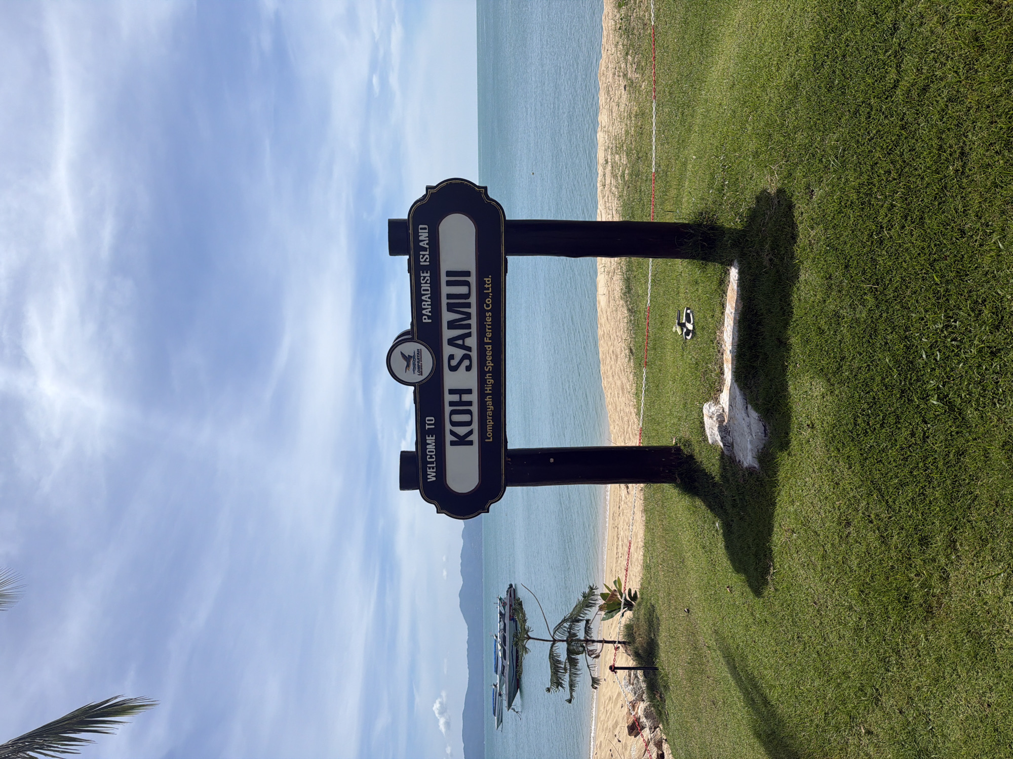 The image depicts a welcome sign for Koh Samui, a paradise island located in Baan Tai, Thailand. The sign, positioned on a grassy area next to a beach, is primarily dark brown with a decorative outline. Inscribed on the sign in bold, white lettering is "KOH SAMUI," along with the phrase "PARADISE ISLAND." Above the island name is a logo, and underneath, it reads "Lomprayah High Speed Ferries Co., Ltd."
Behind the sign, the scene transitions to a sandy beach that meets the ocean. Several boats are anchored in the water, their forms visible against the horizon. The sky above is predominantly blue, speckled with wispy clouds.
Adjacent to the sign and the beach is an expanse of green grass. Near the sign, two sandals rest on the ground. A small palm tree adds a tropical element to the foreground.