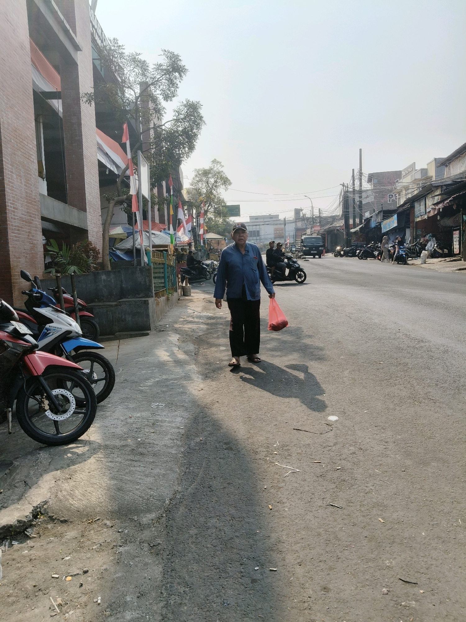 The image depicts a street scene in South Tangerang, Indonesia, on a bright, sunny day with a slightly hazy sky. The perspective is from the street level, looking towards the right side of the road.
In the foreground, on the left side of the image, is the sidewalk and the side of a brick building with horizontal concrete elements. Several motorcycles are parked along the sidewalk. From front to back, a red motorcycle is prominently visible, followed by a blue and white motorcycle, and then a red motorcycle partially obscured.
On the street, a person is walking towards the viewer from the right side of the image. This individual is wearing dark pants, a blue collared shirt, and a baseball cap. They are carrying a translucent pink plastic bag filled with red items in their right hand. Their shadow is cast on the asphalt behind them.
The street itself is paved with asphalt and appears to be a busy thoroughfare with other vehicles visible in the distance, including more motorcycles and a dark van. The right side of the street features a row of buildings with various awnings and signs, suggesting a commercial or residential area. Indonesian flags are visible on poles along the sidewalk near the building on the left.
The overall environment appears to be an urban setting, likely a commercial street with pedestrian and vehicular traffic. The weather is clear, and the sun is casting distinct shadows.