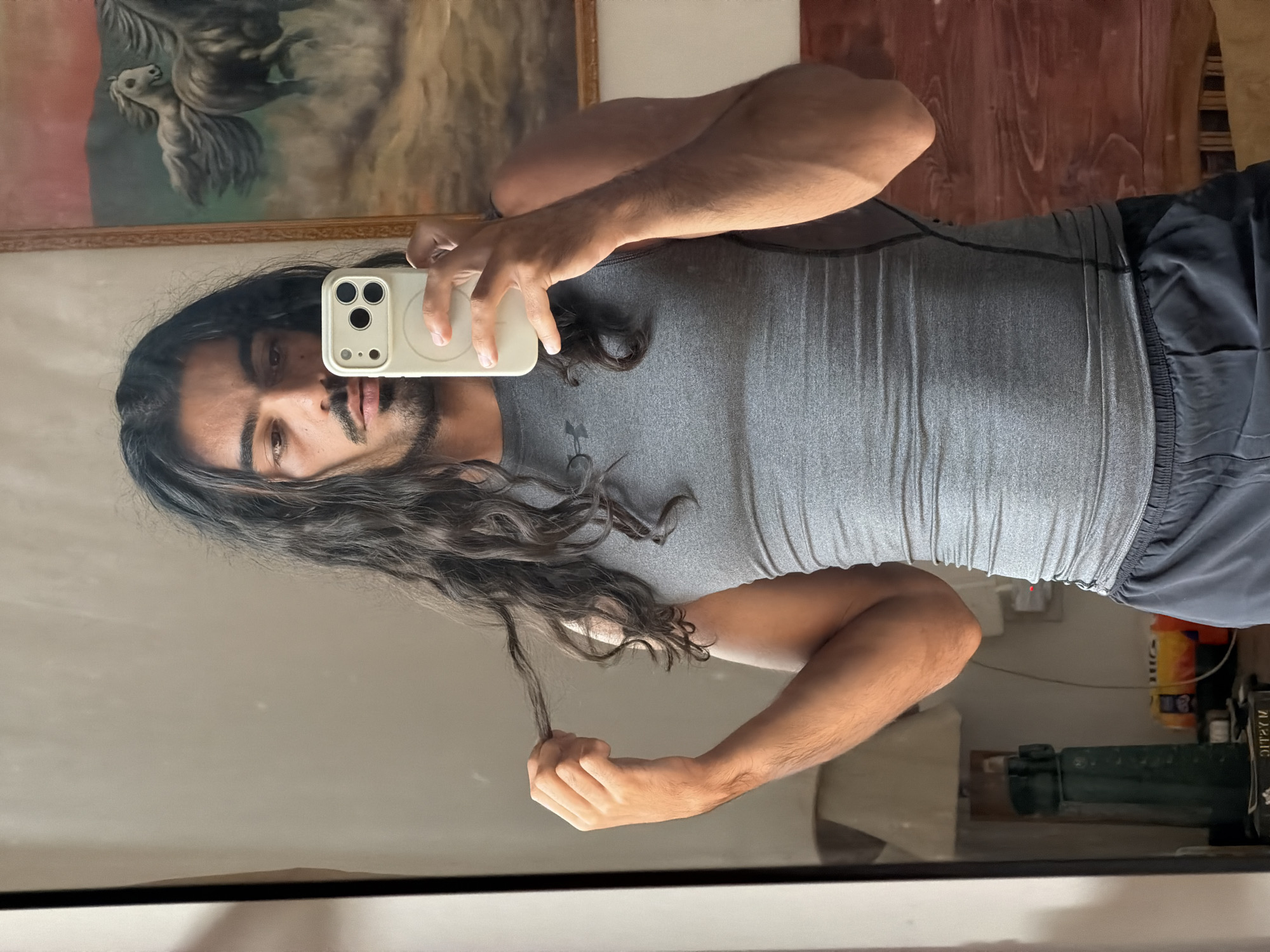 A man with long, dark curly hair is taking a selfie in a mirror. He is wearing a gray Under Armour athletic shirt and dark shorts. He is holding an iPhone with a beige case. In the background, there is a painting of horses. The location appears to be in a bathroom in North Kuta, Indonesia. This is indicated by the presence of cleaning supplies and a green metal scale partially visible in the mirror.