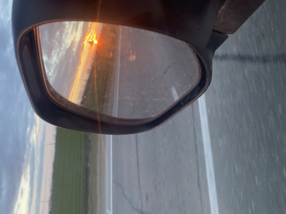 The image displays the black side-view mirror of a vehicle, angled from the right side of the frame, against a rural or semi-rural backdrop in the city of Prostorny, Russia. The mirror's surface appears dusty or smudged and reflects a vivid sunset or sunrise. Within the reflection, the sun, a bright orange circle, is low on the horizon, casting strong orange and yellow light across the sky and clouds. A dark, possibly conical silhouette is partially visible against the sun's glare in the mirror. Outside the mirror, on the left, a grey paved road with a single white edge line is visible, showing minor surface cracks. Beyond the road, a wide expanse of green field or cultivated land stretches to the horizon under a cloudy sky with shades of light blue and grey. No individuals are present in the scene, and no specific actions or interactions are depicted.