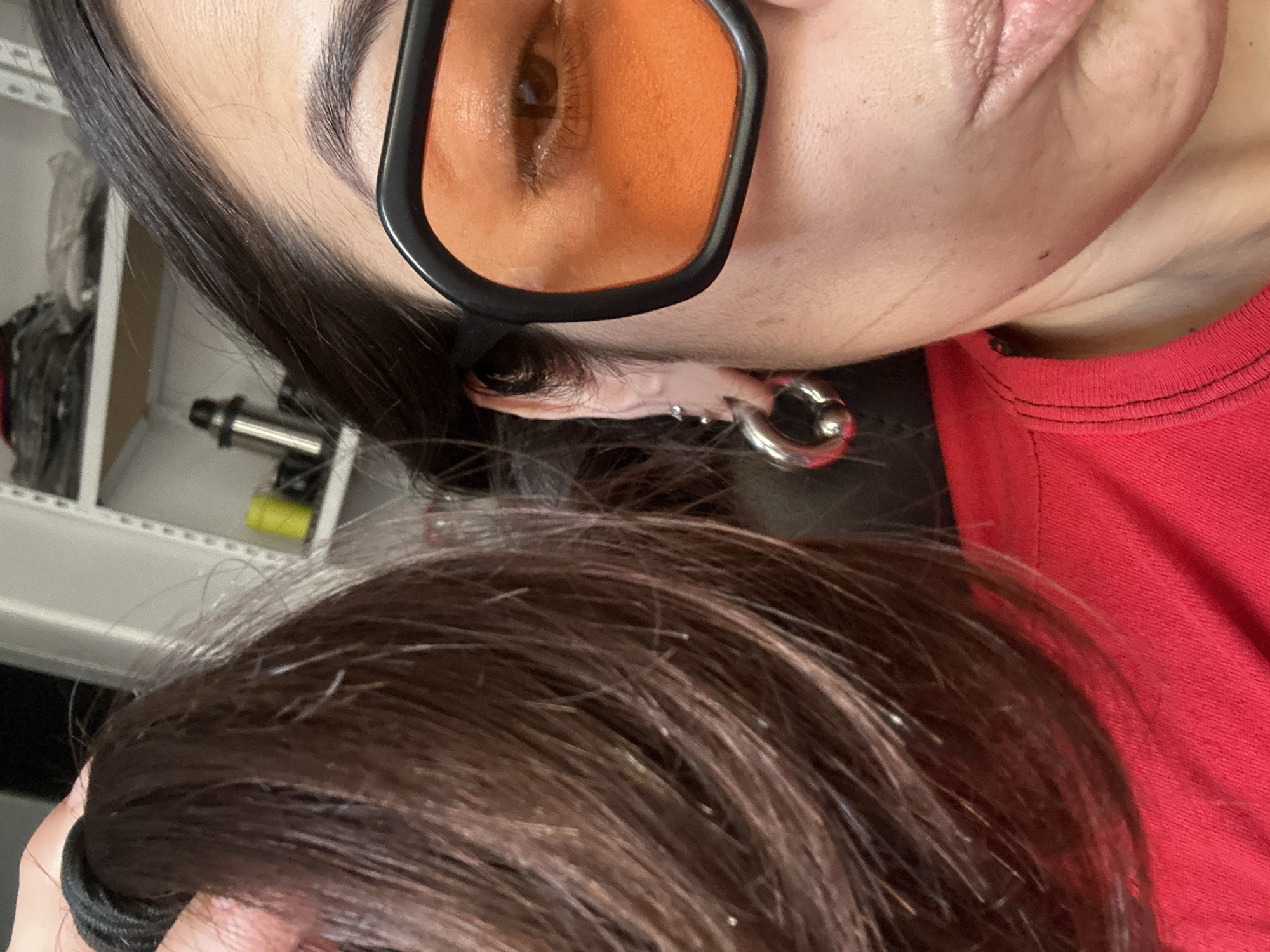 A close-up, tilted perspective features a person wearing orange-tinted glasses and a red shirt. The person has dark hair and an earring. A second person with brown hair is partially visible in the lower portion of the frame. The background includes a shelf. There are no indications of the location being Buenos Aires, Argentina.