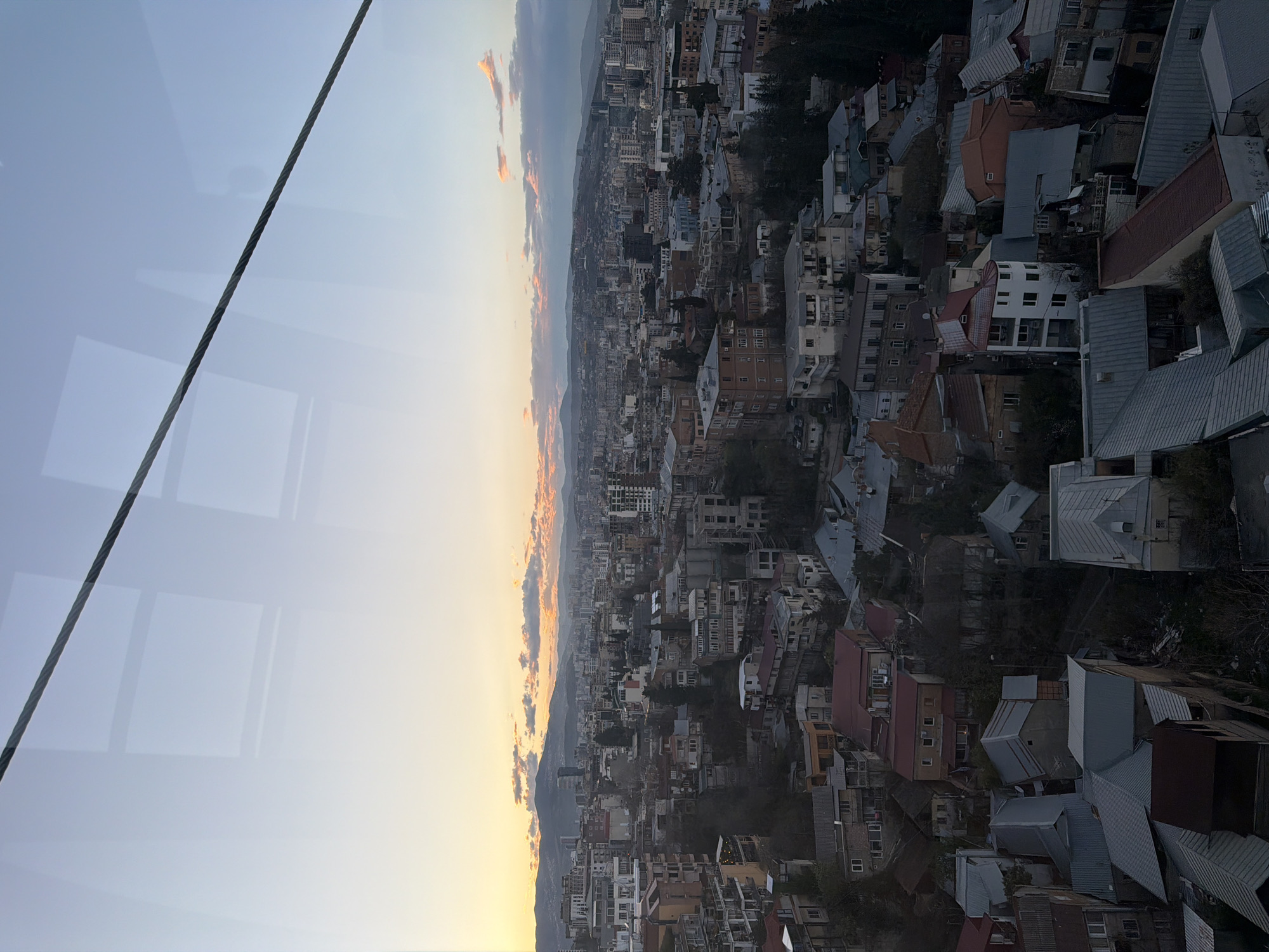 The image presents an aerial view of Tbilisi, Georgia, at dusk or dawn. The cityscape consists of densely packed buildings, predominantly low to mid-rise, with varying architectural styles and roof colors ranging from red and brown to gray. A distant mountain range is faintly visible on the horizon. The sky transitions from a pale blue at the top to a warm, golden hue near the horizon, where the sun is likely setting or rising, with scattered clouds tinged with orange. A steel cable is visible in the upper left corner, suggesting the photo was taken from a cable car or similar elevated structure. The overall environment conveys a sense of urban density within a landscape defined by the surrounding mountains.