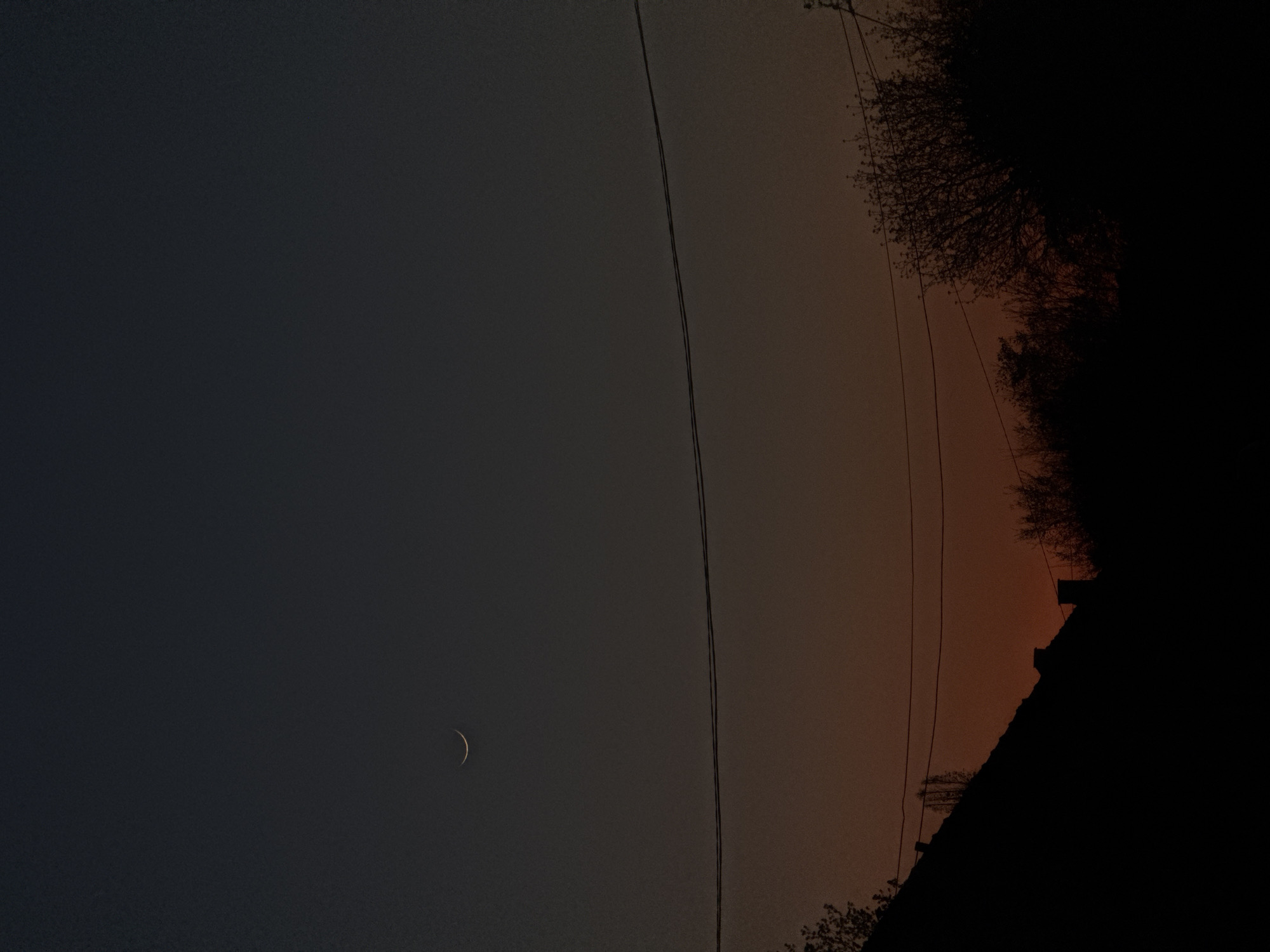The image shows a crescent moon in the sky. Two electrical wires run vertically through the middle of the frame. The sky transitions from a dark blue on the left to a reddish-orange hue on the right. Silhouetted trees and a building are visible on the right edge of the image. The image was taken at night. There is no evidence of the location being Lehchevo, Bulgaria. No people are visible.