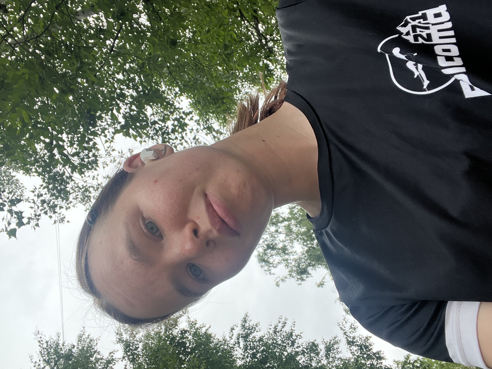 A low-angle, eye-level selfie captures a young Caucasian female looking directly at the camera. The scene is set outdoors in Chaikovsky, Russia, characterized by an overcast sky and abundant green foliage from deciduous trees filling the background.
The subject has light brown hair, pulled back, with some loose strands framing her face and a reddish-brown streak visible near her neck. Her eyes are blue-grey, fixed on the viewer, and her expression is neutral. Her skin has a fair complexion, showing visible pores and minor blemishes on her forehead, left cheek (viewer's right), and neck. She wears a white wireless earbud in her right ear (viewer's left) and two small, silver-toned stud earrings in her left earlobe and helix (viewer's right). Her lips are naturally pink and slightly parted.
She is wearing a black, short-sleeved, crew-neck t-shirt made of a textured athletic fabric. The left sleeve (viewer's right) features a white cuff. On the lower right chest of the shirt, a white graphic logo is visible. This logo depicts stylized mountains on the right, with the number "271" integrated into one of the peaks. To the left, a figure appearing to be a runner or skier is positioned within a shield-like outline. Below these elements, the word "Picoma" is printed, with the 'P' and part of the 'i' clearly visible.
The background primarily consists of a light grey, overcast sky, indicating cloudy conditions. Numerous green leaves and branches of trees frame the subject, suggesting a park or wooded area. A light-barked tree trunk is partially visible on the upper right side of the frame. Faint, thin lines, possibly power lines, stretch across the upper left sky.