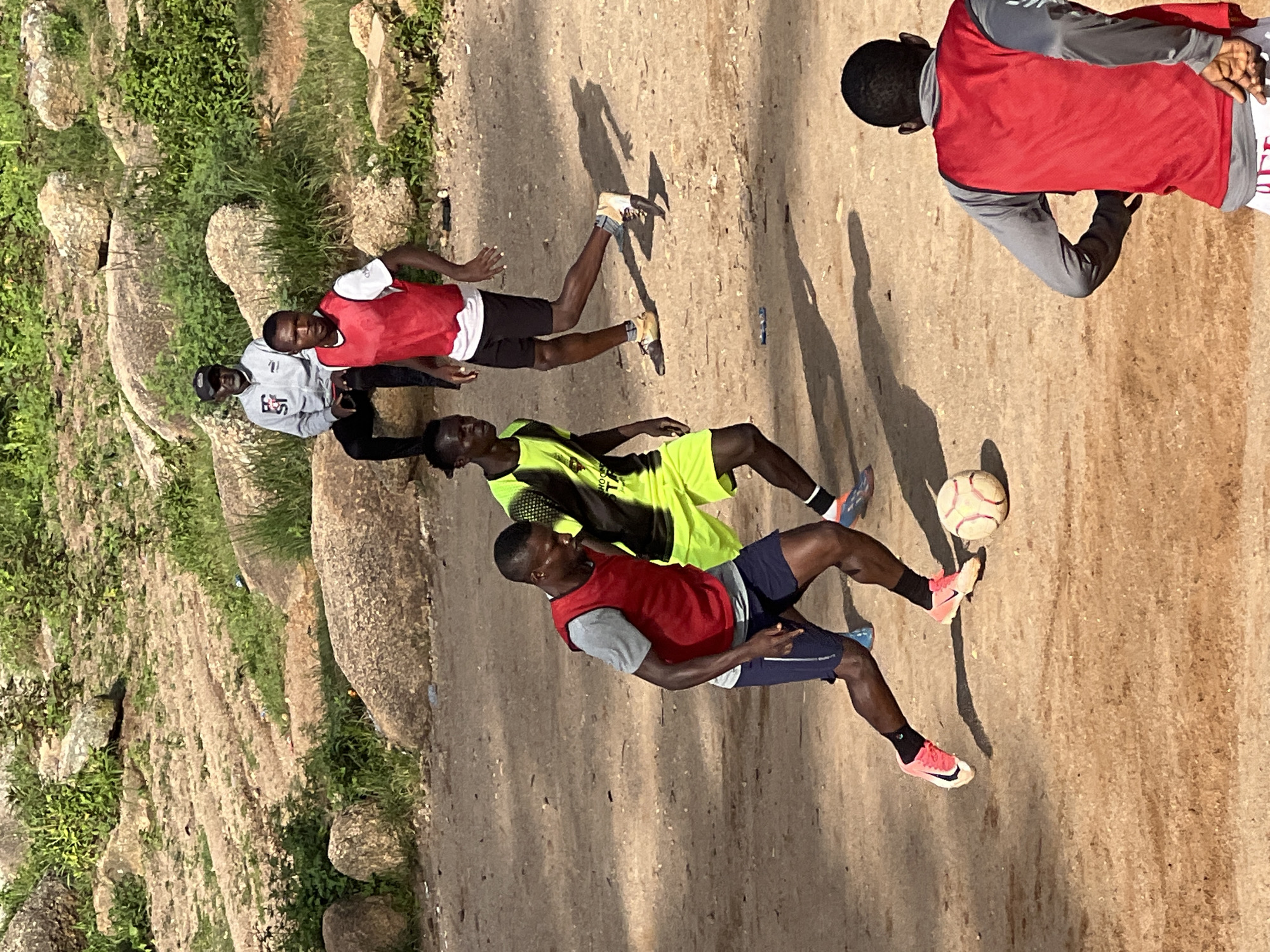 In Jos, Nigeria, on a dirt field with rocky terrain and sparse green vegetation, a group of young men are engaged in a football game. The ground is composed of reddish-brown dirt, with large, weathered rocks scattered throughout the landscape. In the foreground, one player in a red vest and blue shorts is dribbling a football, his body angled forward in motion. To his left, another player in a neon yellow and black jersey is also in pursuit of the ball. Further in the background, a man in a grey jacket and black cap sits on a rock, observing the game. Another player in a red vest and white shirt stands to the right, facing away from the camera. The atmosphere appears casual and recreational, typical of informal football matches played in open spaces in Jos.