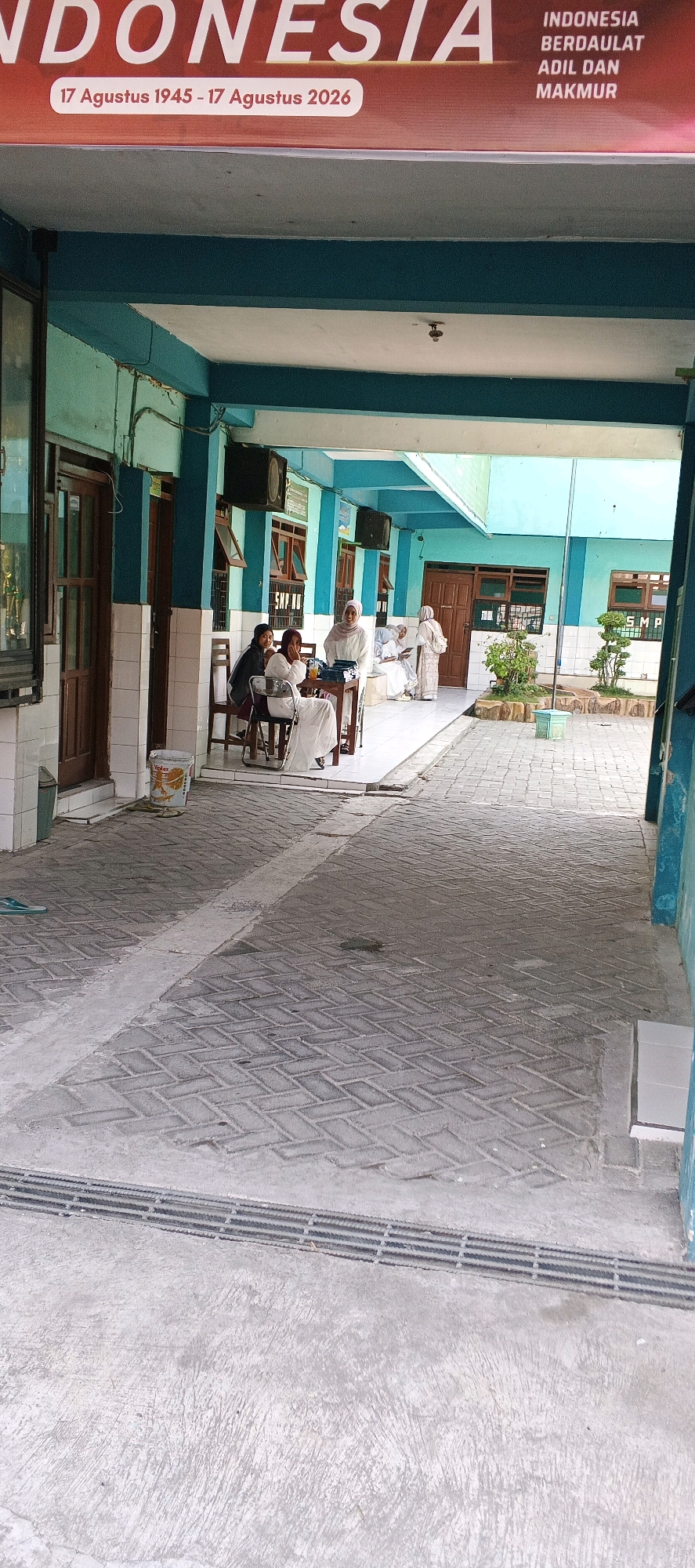The image shows a school hallway with a group of people. A banner at the top reads "INDONESIA BERDAULAT ADIL DAN MAKMUR" and "17 Agustus 1945 - 17 Agustus 2026". There are several doors on the left and right sides of the hallway. In the middle of the hallway, there are three women sitting at a table, and two more women standing behind them. One woman is holding a glass with a straw. The floor is paved with interlocking bricks. A trash can is visible near the left wall. The school appears to be named SMP. The scene is outdoors, likely in a tropical climate due to the presence of greenery and the style of the building.