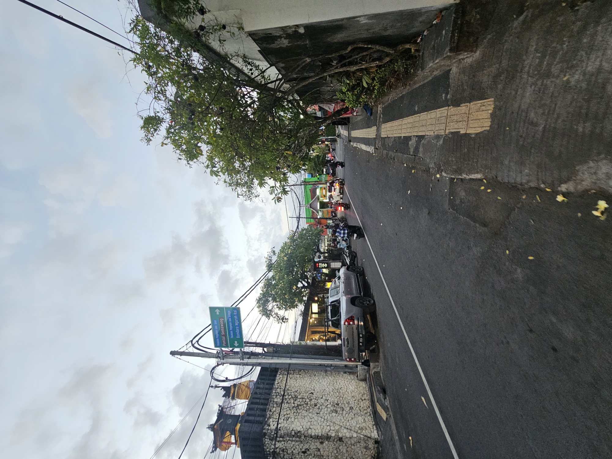 This is a street-level, wide-angle shot of a road in Denpasar, Indonesia, likely during the late afternoon or early evening given the overcast sky and the lighting conditions. The scene depicts a moderately busy street with traffic, including motorcycles and at least one car.
On the left side of the road, there is a stone wall and a pole with two green directional signs. The upper sign reads "Pelabuhan Umpal Sidur" with an arrow pointing right. Below it, a sign reads "Pantai Semawang" with an arrow pointing right. The lowest sign reads "Bandara Ngurah Rai" with an arrow pointing left. A silver pickup truck is parked on the side of the road in front of a low-rise building with large windows, possibly a shop or restaurant. A motorcycle is parked behind the pickup.
The road itself is paved asphalt with a white line marking the edge of the lane. There are several vehicles and motorcycles on the road, moving away from the camera. The right side of the road features a sidewalk with tactile paving for visually impaired individuals, adjacent to a wall covered in vines. Trees with green foliage line the right side of the sidewalk.
The sky is overcast with scattered clouds. The overall impression is of a typical urban street scene in a tropical environment, with a mix of residential and commercial elements, and active local transportation.