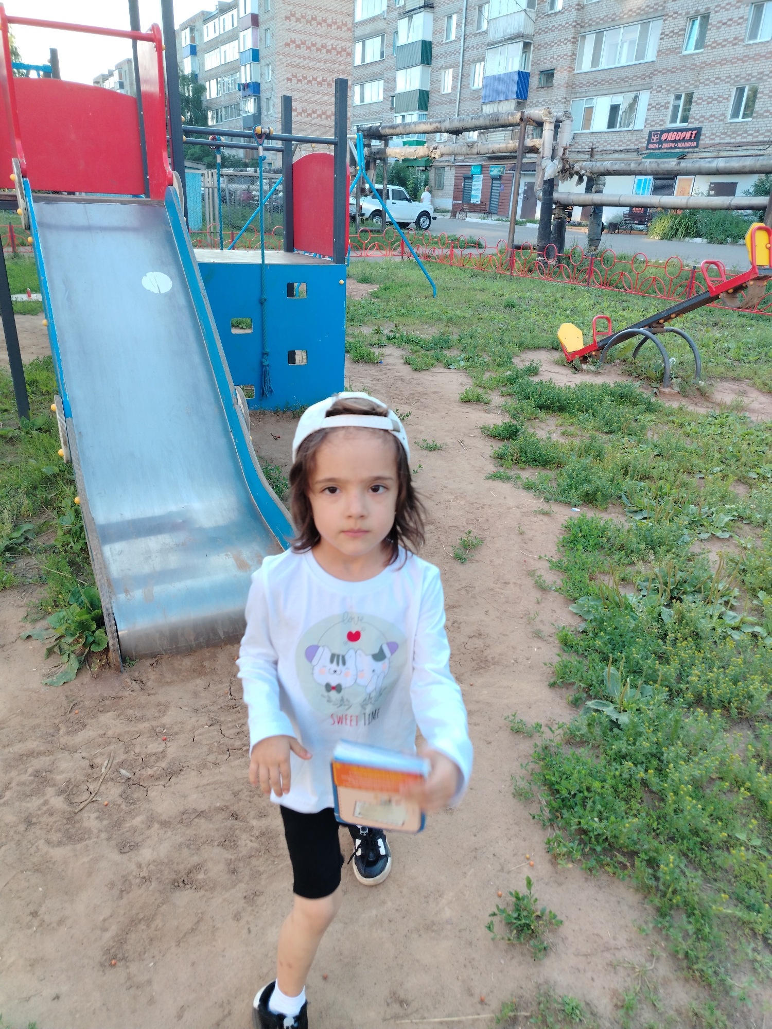A young girl with dark, shoulder-length hair and a white baseball cap stands in a playground in Yanaul, Russia. She is wearing a long-sleeved white shirt with a graphic of two cartoon cats and the words "SWEET TIME," black shorts, white socks, and black and white sneakers. She is holding a small orange and blue object, possibly a book or toy, in her right hand.
The playground features a metal slide with a red platform and blue structural elements. To the right of the slide, a seesaw with yellow and red seats is visible. The ground is a mixture of dirt and sparse green vegetation. In the background, a multi-story brick apartment building is visible, with a street and a white car parked in front of a commercial building with signs. Heating pipes run horizontally across the middle ground. The scene appears to be captured during daylight hours.