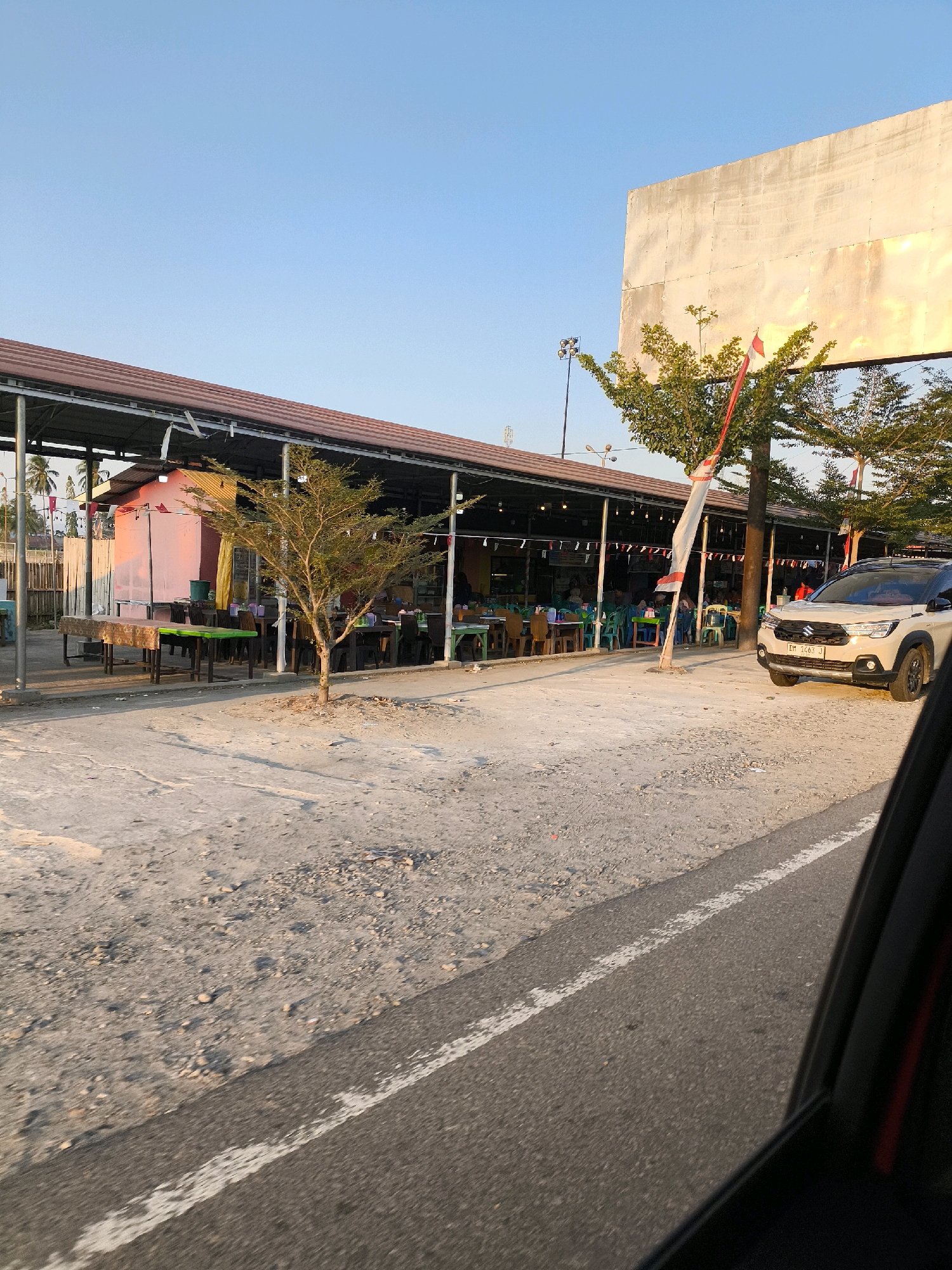 The image captures an outdoor dining area in Tenilo, Indonesia, under a clear sky with the late afternoon sun casting a warm glow. The scene is dominated by a long, open-sided structure with a reddish-brown roof, supported by metal posts. Beneath this structure, numerous tables and chairs are arranged, some covered with checkered cloths. The ground is a mix of gravel and sand.
To the right, a light-colored SUV is parked. In the background, a large, blank billboard-like structure stands tall. Indonesian flags, red and white, are visible, one prominently displayed on a pole near a tree. The environment suggests a roadside eatery or a market area. The perspective is from a vehicle moving along a paved road, indicated by the visible road surface and white lane markings.