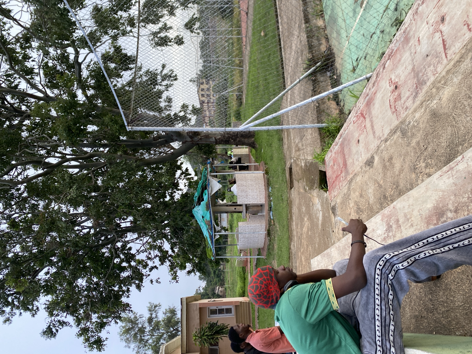The image depicts a scene with several people seated on concrete bleachers, with a lush green tree to the left. The people appear to be casually dressed, with one individual wearing a green t-shirt and a red and black patterned beanie, and another in a grey tracksuit with decorative stripes. They are looking upwards and to the right, possibly at an event or a point of interest not fully captured in the frame. The setting appears to be an outdoor recreational area, perhaps a sports field or a park, with a chain-link fence and a green surface, possibly a tennis court or a playing field, visible in the background. A small, open-air structure with a blue tarp roof is also present, suggesting a shelter or vendor stall. The overall lighting suggests it might be daytime, with overcast or diffused light. The presence of buildings in the distance hints at an urban or semi-urban environment within Jos, Nigeria. No text is visible in the image.