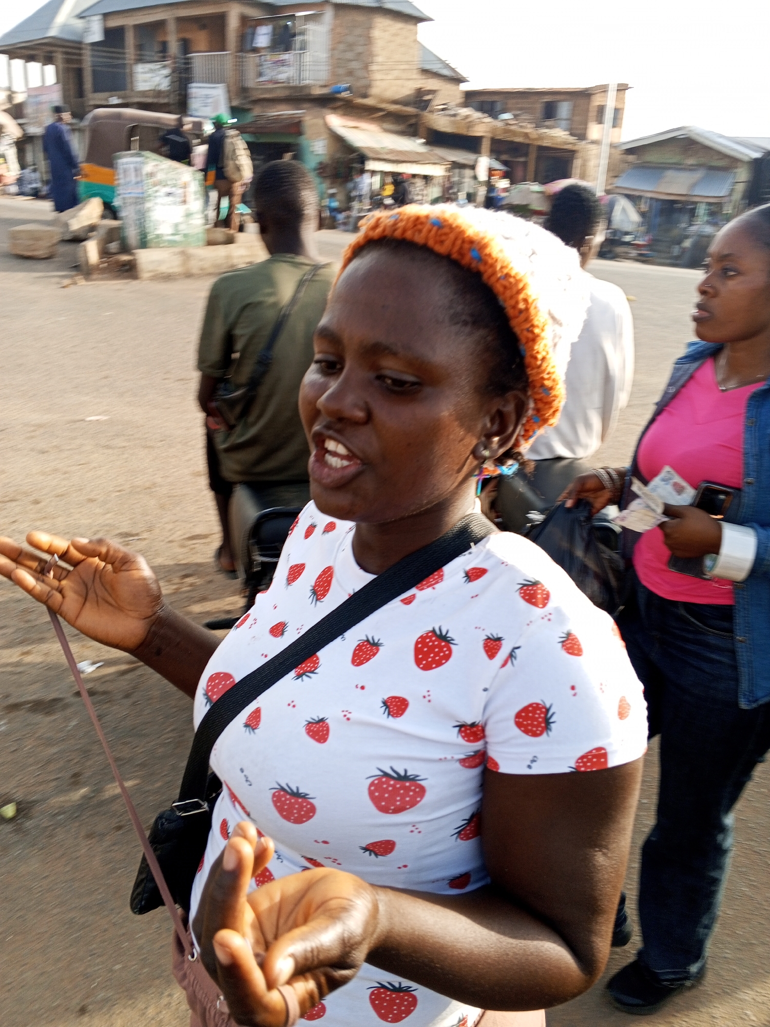 The image depicts a woman with dark skin and a white t-shirt covered with strawberry prints, wearing a white and orange knit cap. She's in mid-sentence, her mouth open as if speaking. She's gesturing with her left hand, holding a thin strap. A black shoulder bag is visible across her body. To her right stands another woman in a pink shirt and denim jacket, holding what appears to be Nigerian currency and a cell phone. Behind them are other people, including one person in a green shirt, and a partially visible motorcycle. In the background are buildings suggesting an urban environment. Based on the prompt, the location is Gwong, Nigeria. The ground appears to be a dirt or concrete surface. An auto rickshaw is visible in the background on the left.