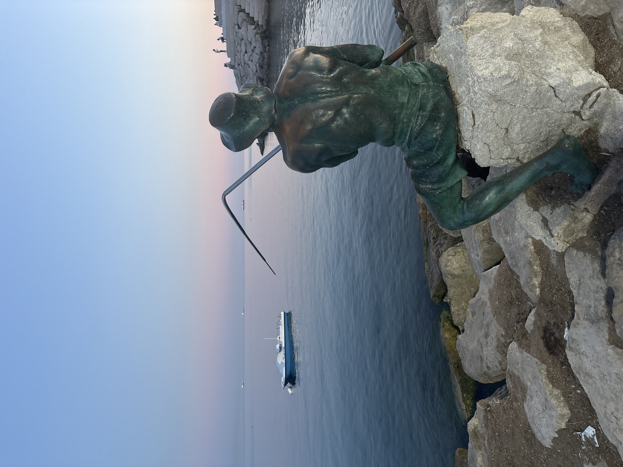A bronze statue of a fisherman is seated on rocks by the sea, facing away from the camera. The fisherman statue is leaning on a fishing rod. The sea is calm and blue. A small blue boat with a white cover is moored in the water. In the background, there is a stone promenade with a few people standing and sitting. The sky is a gradient of blue and pink, indicating dusk or dawn. The scene is located in Durrës, Albania.